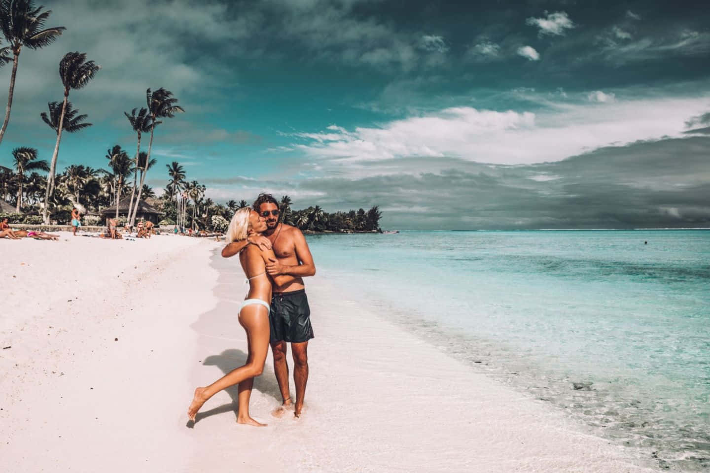 Bora Bora Beach 桌布 壁紙 — 一對情侶在原始白色沙灘上擁抱，背景是戲劇性的多雲天空、碧綠海水和棕櫚樹。
