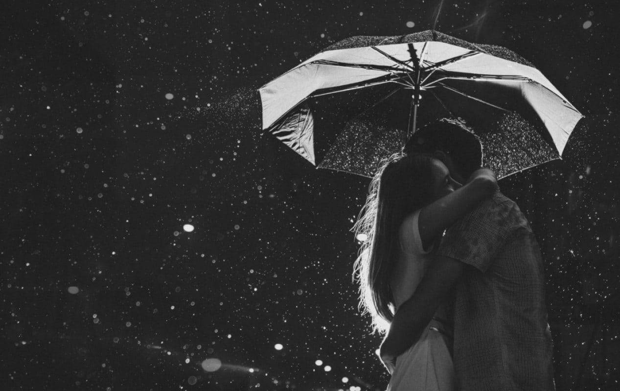 情侶 桌布 壁紙 — 一對情侶在單一把雨傘下擁抱，雨滴在漆黑的夜空下墜落，一張溫柔的黑白照片。