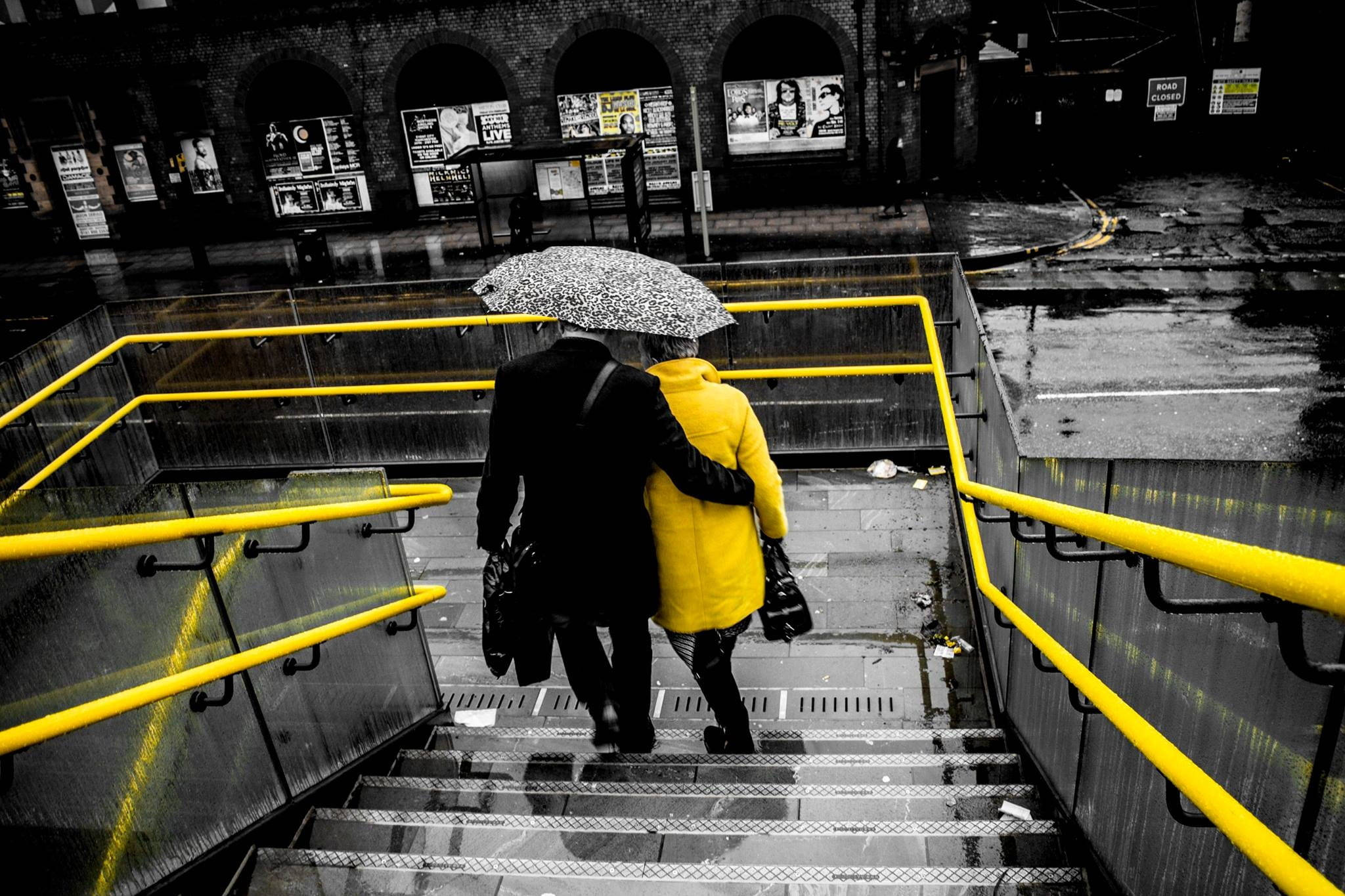 Couple 桌布 壁紙 — 一對情侶在單色城市場景中，撐著圖案雨傘走下濕漉漉的樓梯，旁邊有鮮黃色的欄杆。