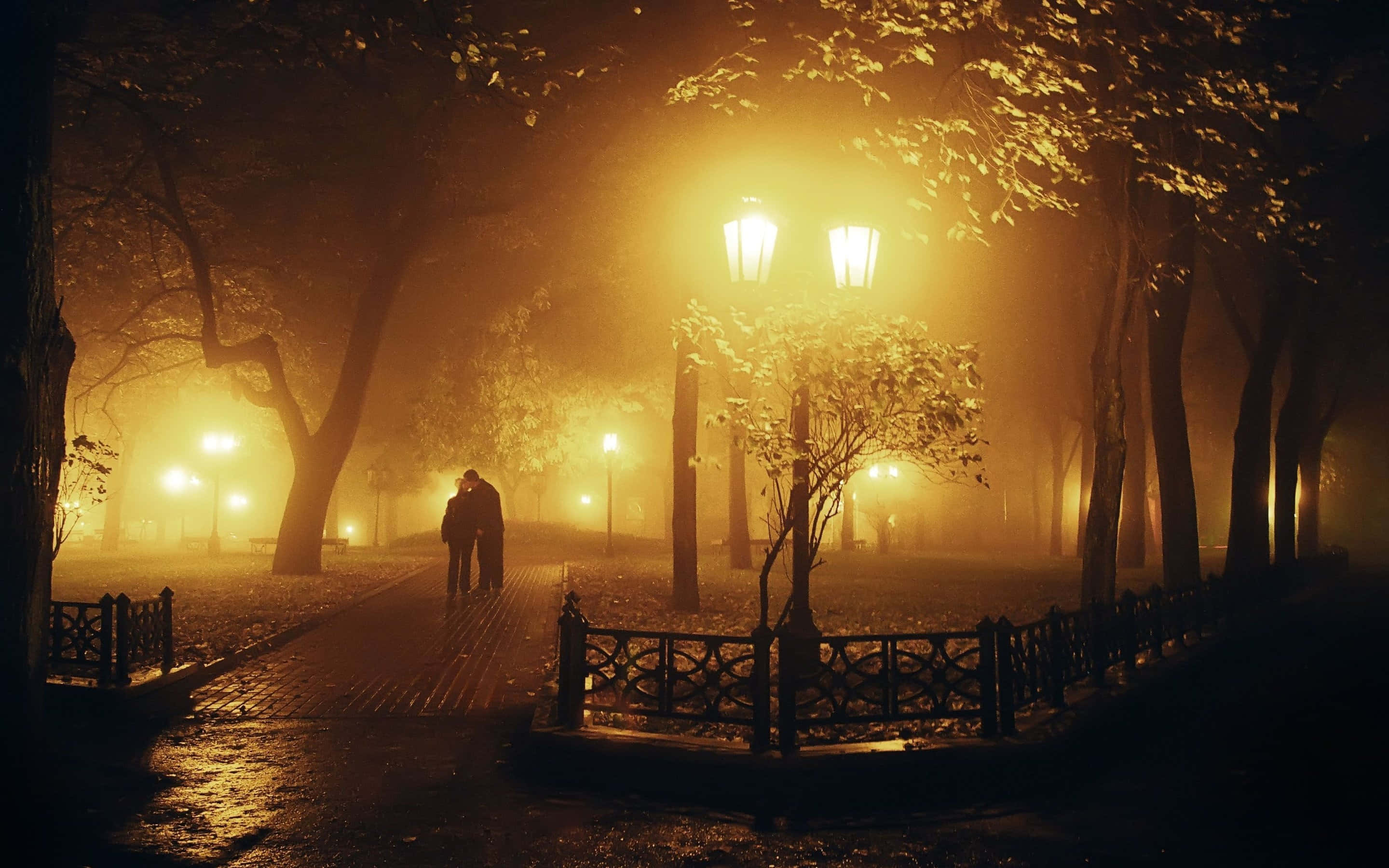 夜晚 桌布 壁紙 — 一對情侶在夜晚手挽手走過霧氣瀰漫的公園小徑，沐浴在復古街燈的溫暖光芒中。