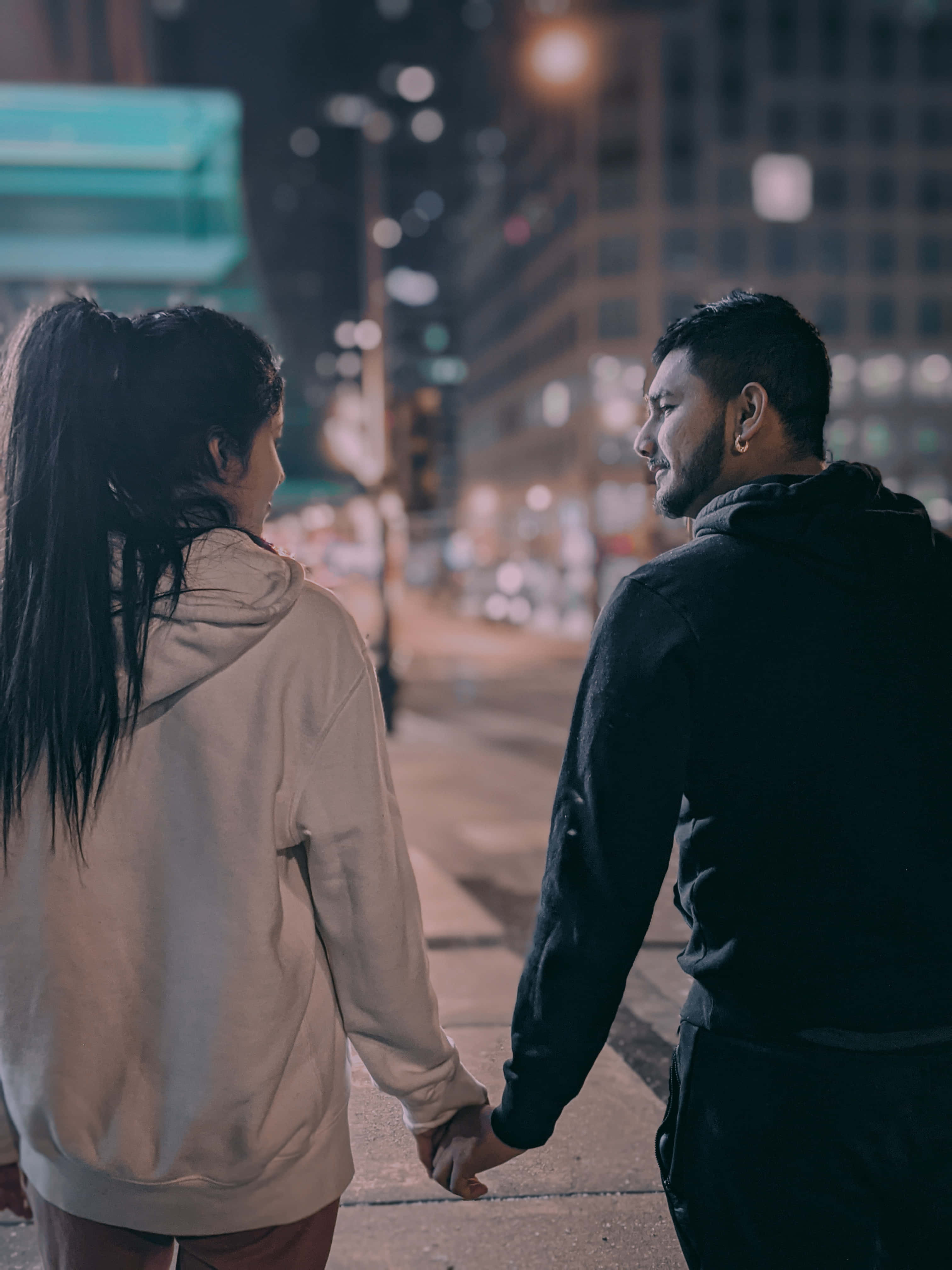 Romance 桌布 壁紙 — 一對情侶在夜晚手牽手走在城市街道上，被溫暖的路燈和身後模糊的建築燈光照亮。