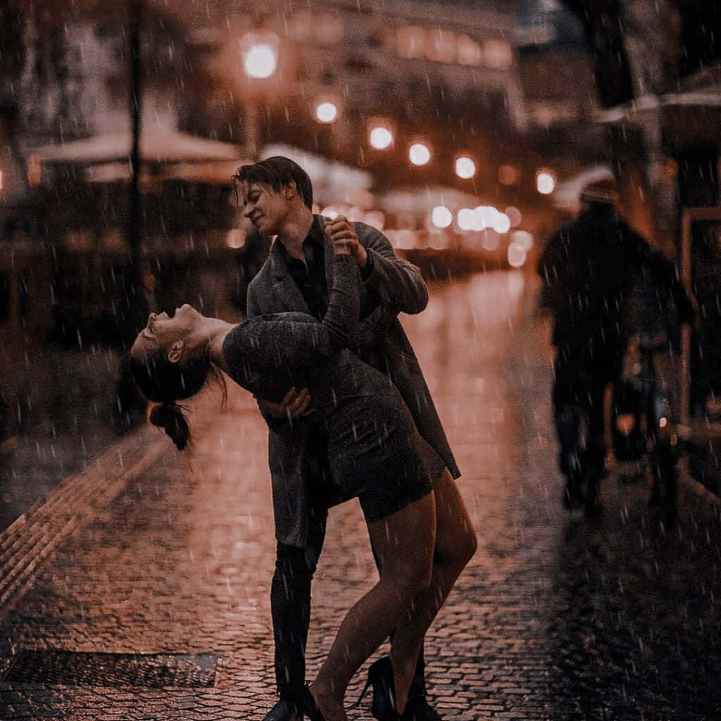 Couple In Rain 桌布 壁紙 — 一對情侶在夜晚溫暖燈光照亮的鵝卵石街道上，在雨中熱情擁抱和跳舞。