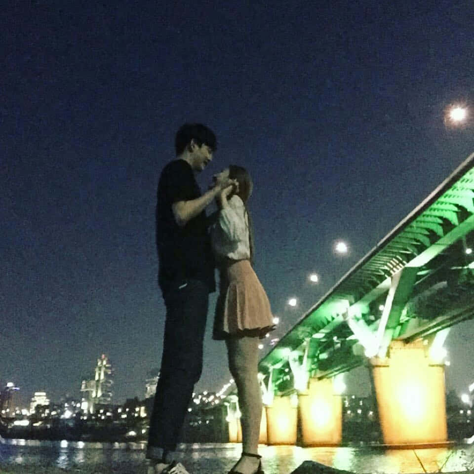 Couples 桌布 壁紙 — 一對情侶在夜晚發光的綠色橋下分享親密時刻，身後的城市燈光倒映在水面上。