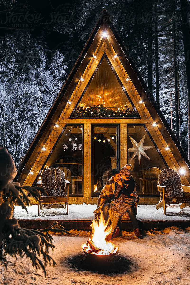 Winter House 桌布 壁紙 — 一對情侶在夜間於被雪覆蓋的A字形小屋外，在發光的火坑旁接吻，周圍是結霜的樹木。