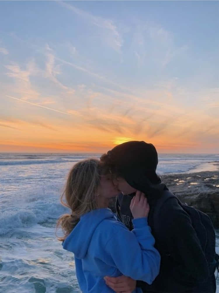 Girlfriend And Boyfriend 桌布 壁紙 — 一對情侶在岩石海灘上親吻，夕陽以火熱的橙色和柔和的藍色色調沉入海洋。