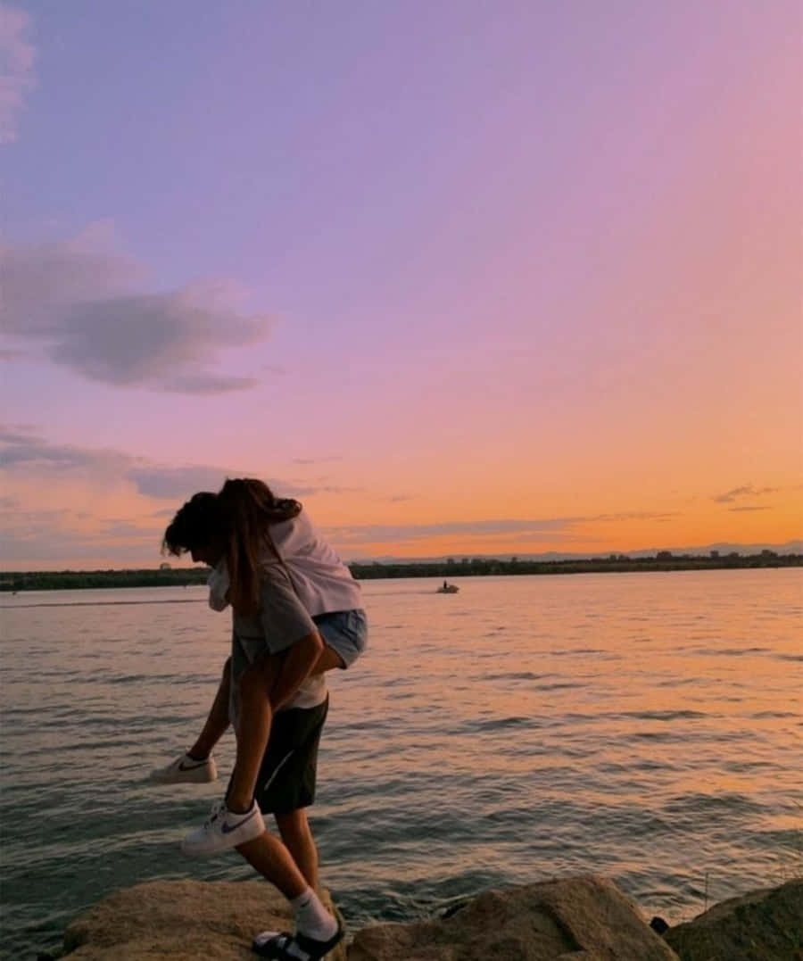 boyfriend 桌布 壁紙 — 一對情侶在岩石湖岸上擁抱，天空燃燒著橙色、粉紅色和紫色的日落色調。