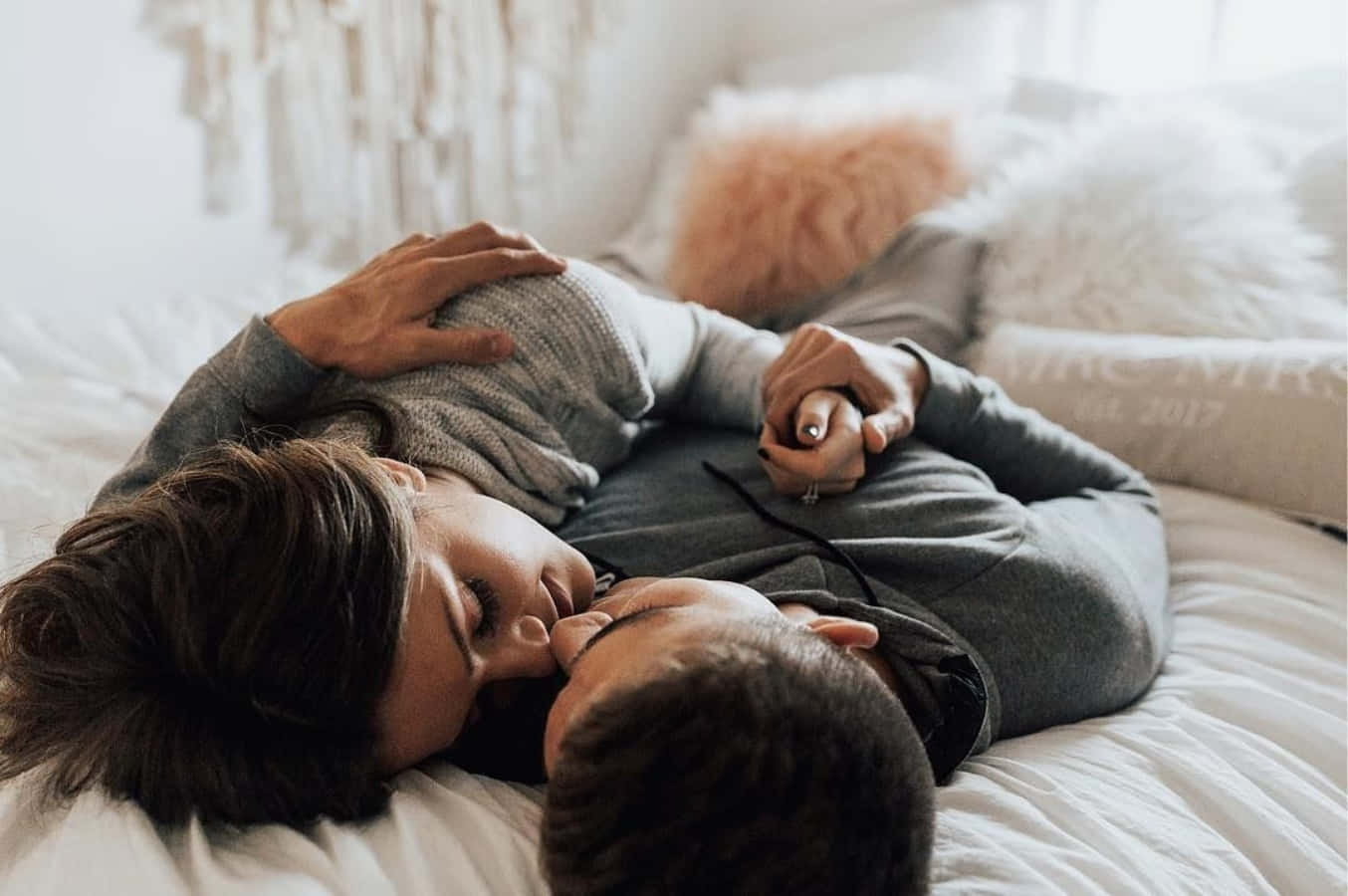 Kissing 桌布 壁紙 — 一對情侶在床上平靜地休息，在柔和自然光下溫柔擁抱，旁邊有 fluffy 枕頭和一個「MRS」枕頭。