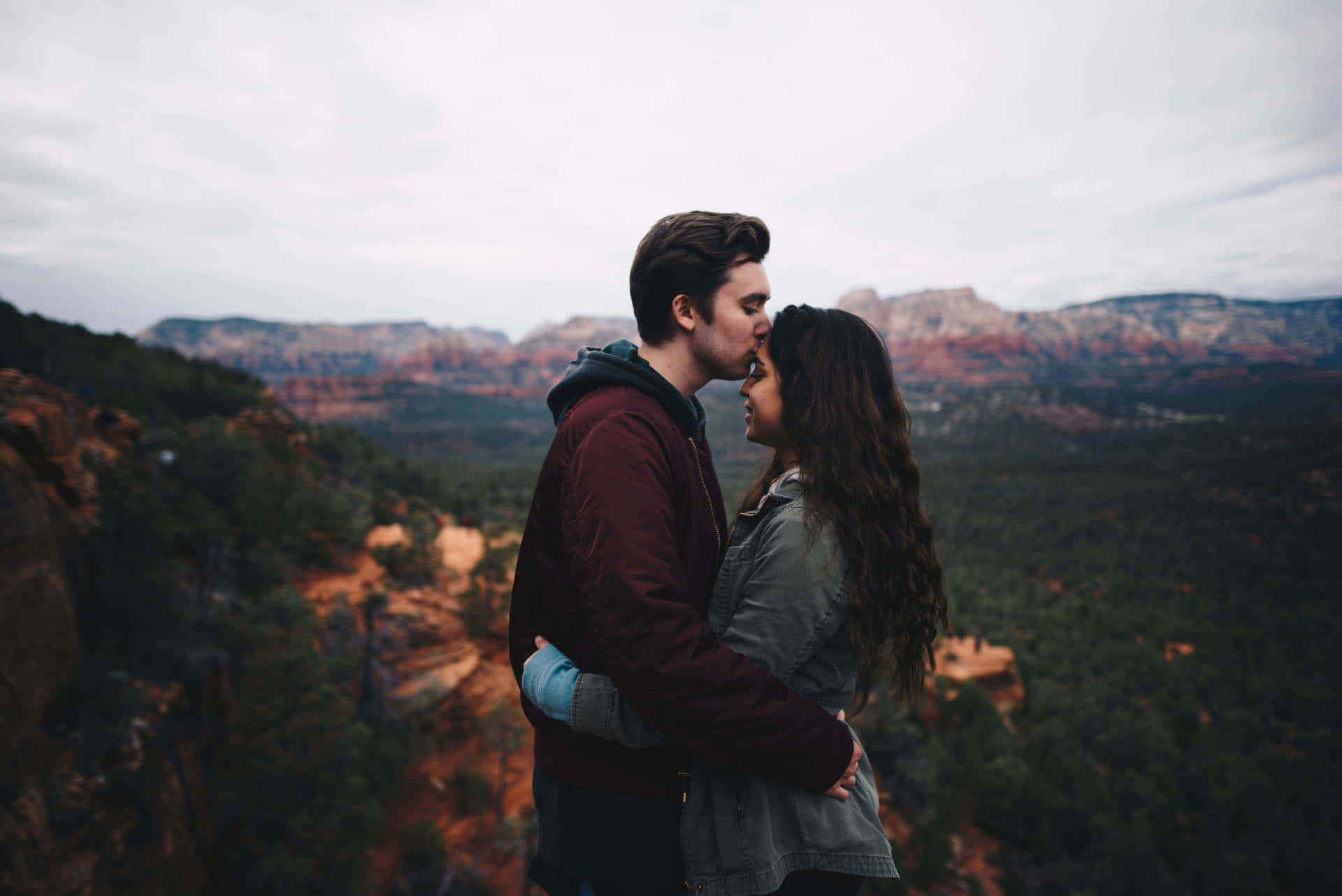 Romantic Love 桌布 壁紙 — 一對情侶在懸崖邊溫柔擁抱，俯瞰陰天下的紅色岩石峽谷。
