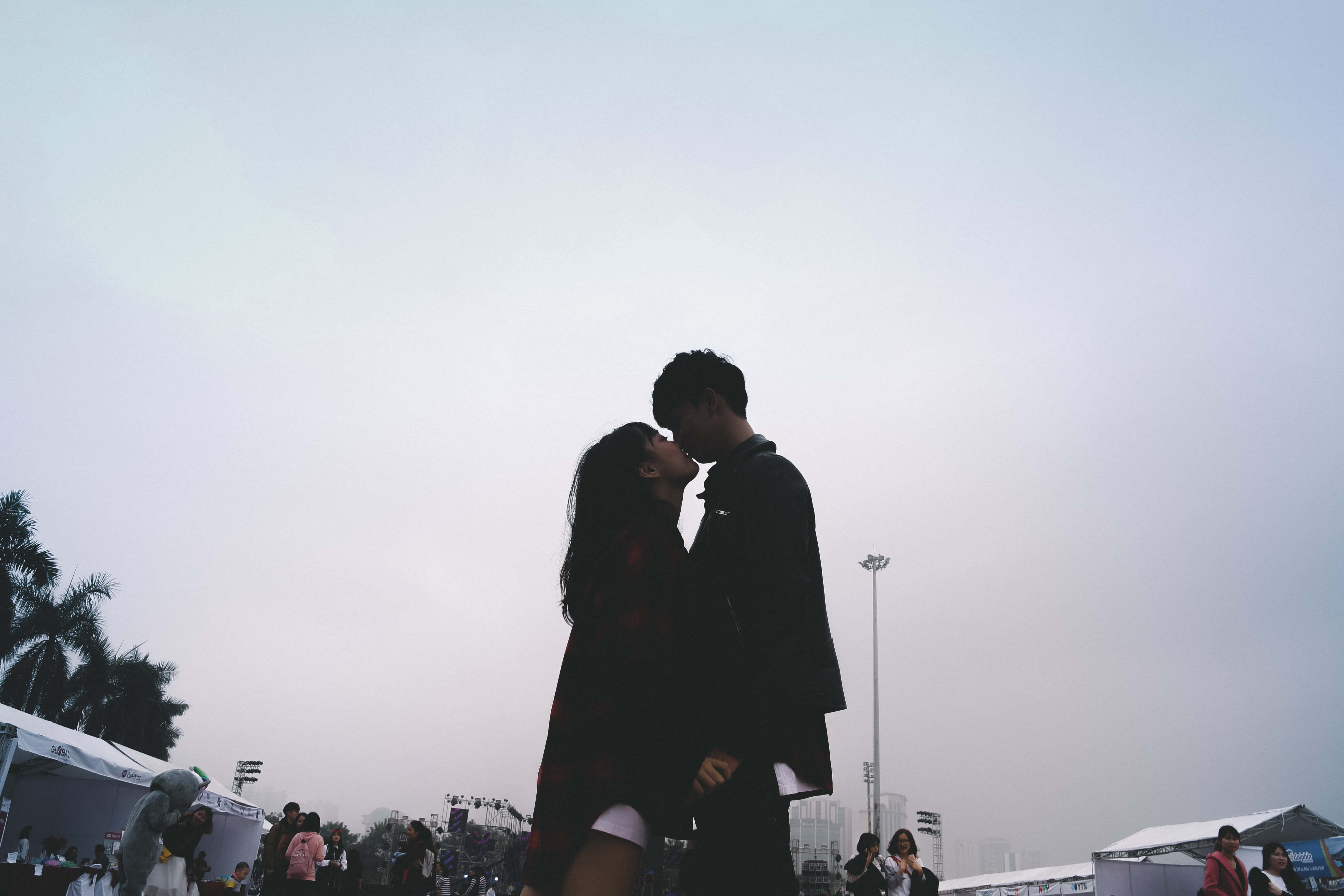 Kiss 桌布 壁紙 — 一對情侶在戶外活動中，以棕櫚樹和帳篷為背景，在朦朧的天空下以剪影接吻。
