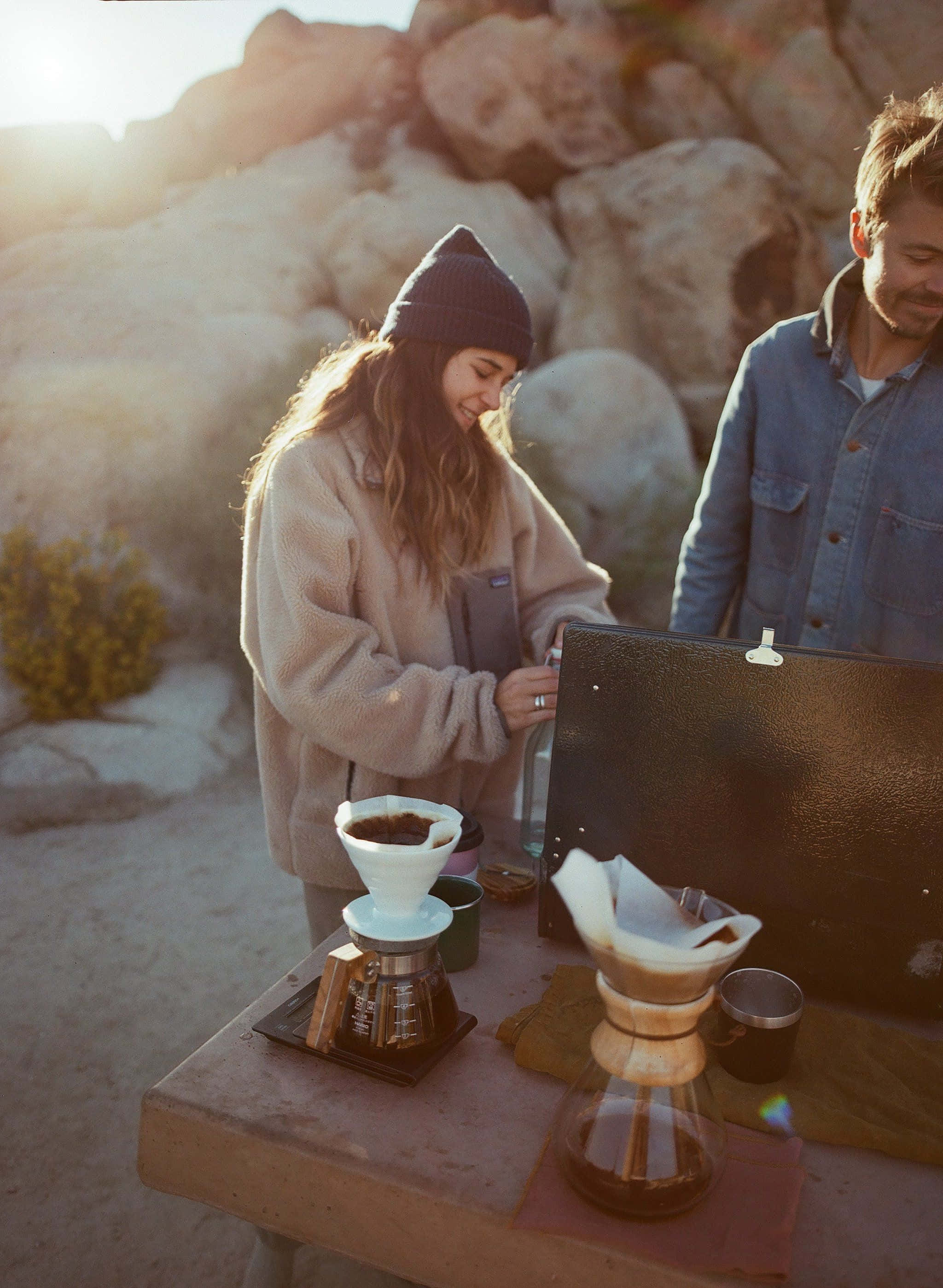 Minimalist Camping 桌布 壁紙 — 一對情侶在日落時分享受戶外手沖咖啡，溫暖的金色光線和岩石沙漠背景。