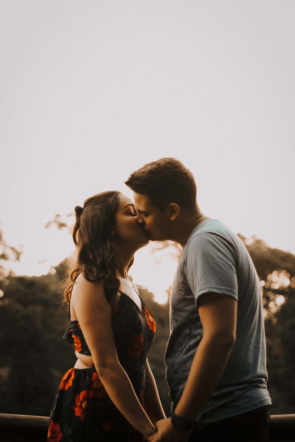 Kissing 桌布 壁紙 — 一對情侶在日落時分享溫柔的親吻，溫暖的金色光芒在他們身後閃耀，樹木在背景中柔和地模糊。