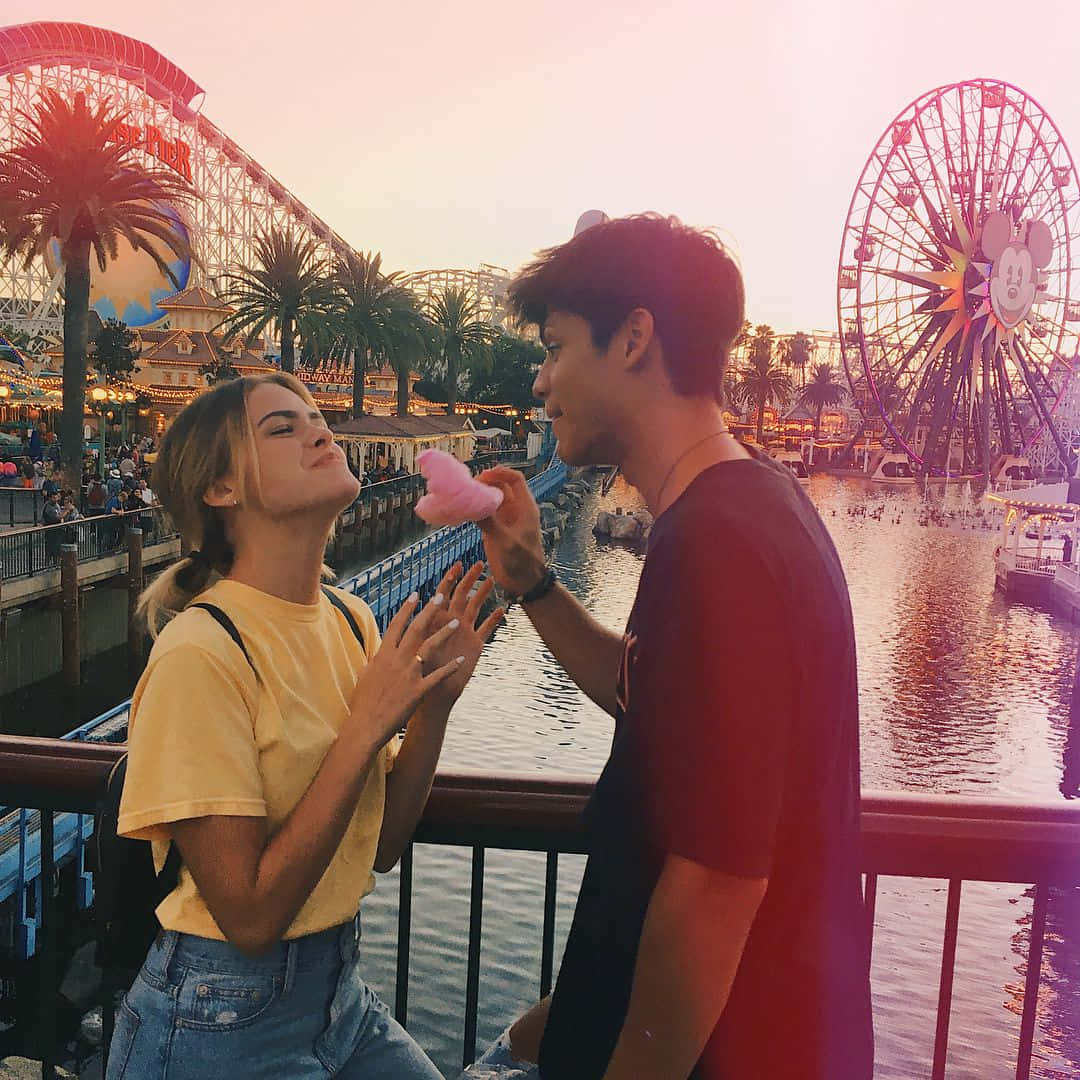 Boyfriend And Girlfriend 桌布 壁紙 — 一對情侶在日落時分享粉紅色棉花糖，背景是充滿活力的遊樂園，有摩天輪和雲霄飛車。