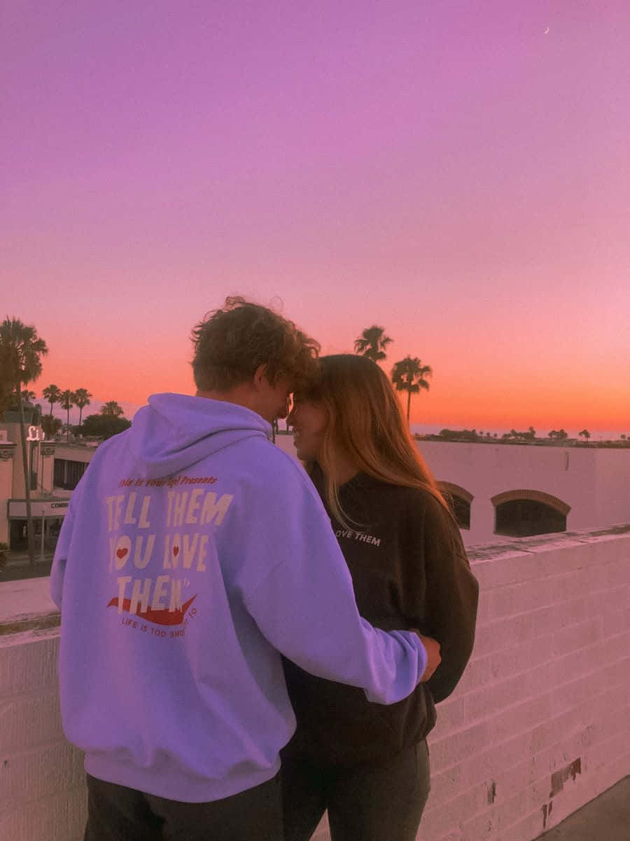 Aesthetic Couple 桌布 壁紙 — 一對情侶在日落時分於屋頂上擁抱，背景有棕櫚樹和粉橙色天空。
