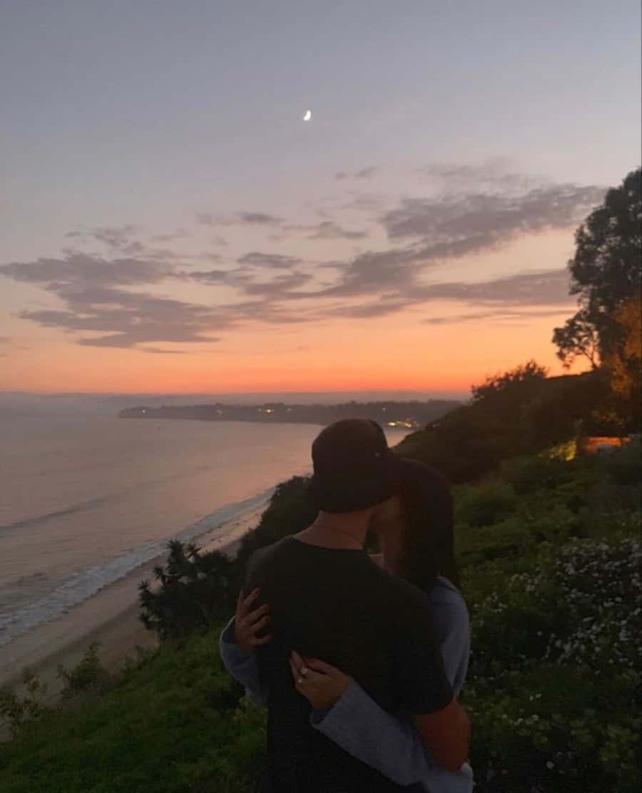 Falling In Love 桌布 壁紙 — 一對情侶在日落時分於懸崖邊相擁，俯瞰海洋，上方有一彎新月。