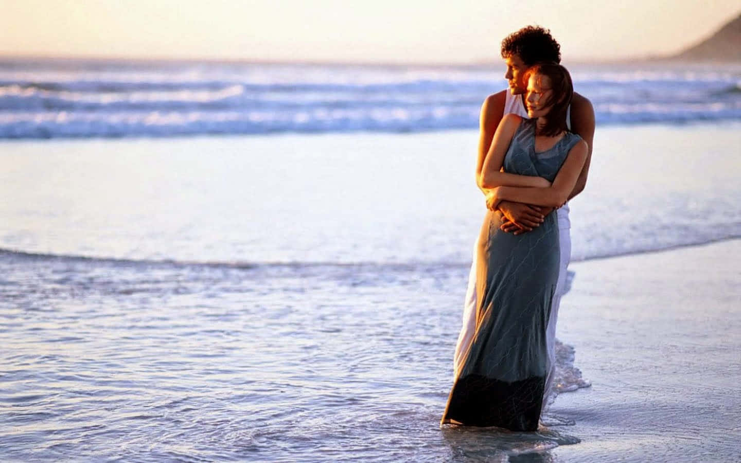 Romantic Love 桌布 壁紙 — 一對情侶在日落時分於淺海水中溫柔擁抱，伴隨著輕柔的波浪和遠山。