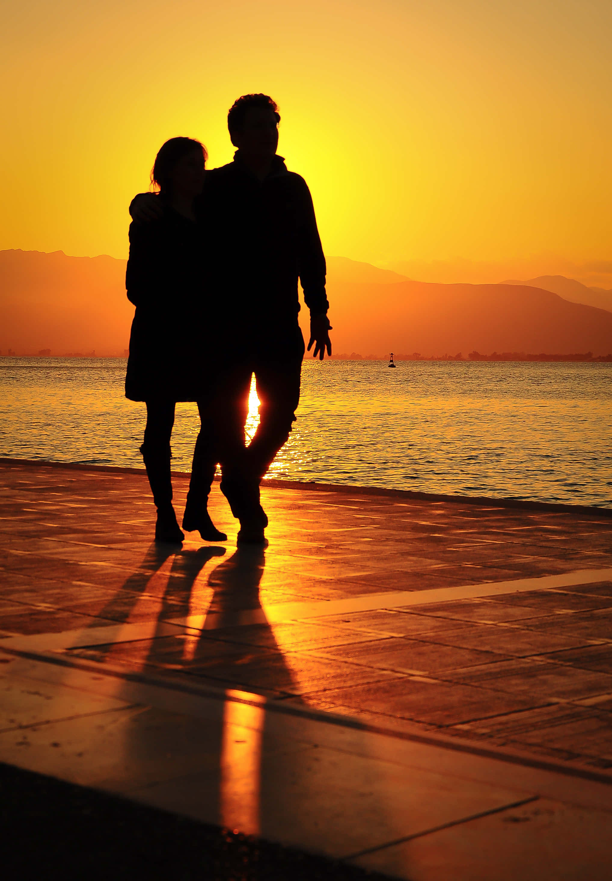 Romance 桌布 壁紙 — 一對情侶在日落時分沿著海濱漫步，他們的剪影在濕漉漉的人行道上投下長長的金色倒影。