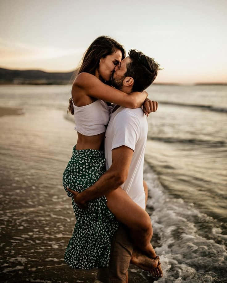 Kissing 桌布 壁紙 — 一對情侶在日落時分的沙灘上親密接吻，溫柔的海浪拍打著他們的腳。