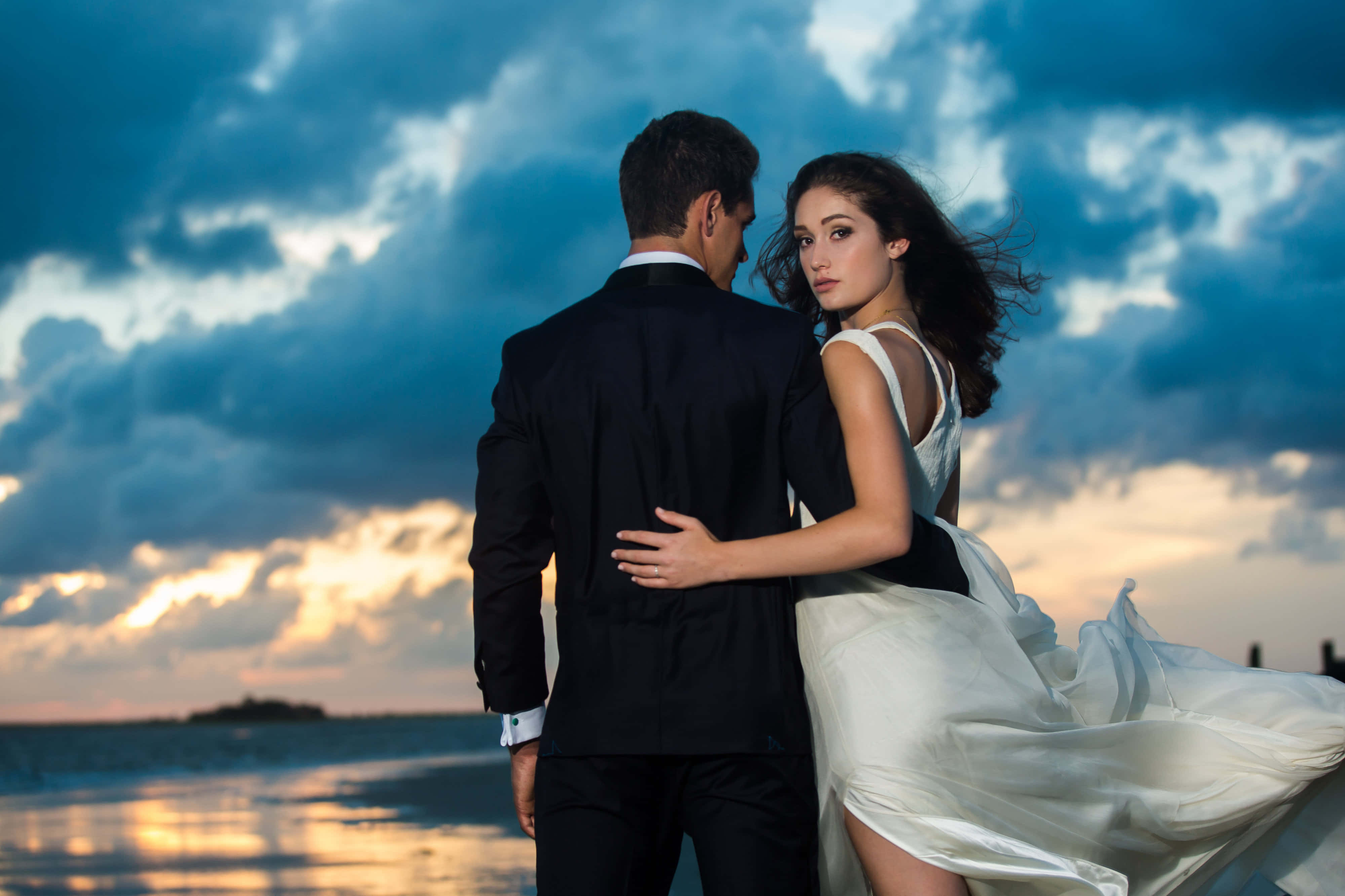 Wedding 桌布 壁紙 — 一對情侶在日落時分的海灘上擁抱，天空有戲劇性的雲彩和金色的水面倒影。
