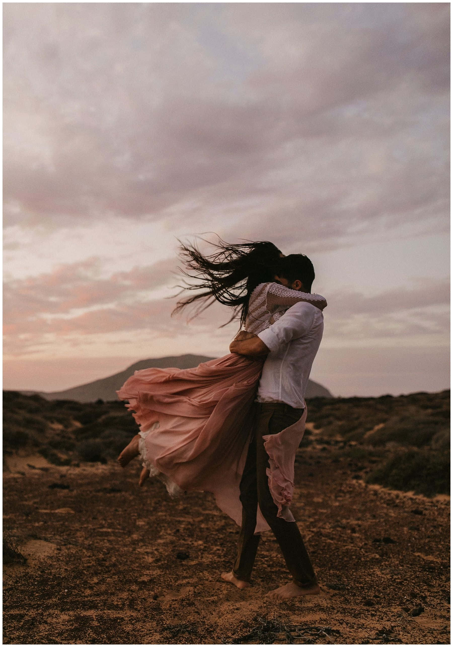 Hugging 桌布 壁紙 — 一對情侶在日落時分的風沙沙漠中熱情擁抱，背景有粉紅色的雲和遠山。