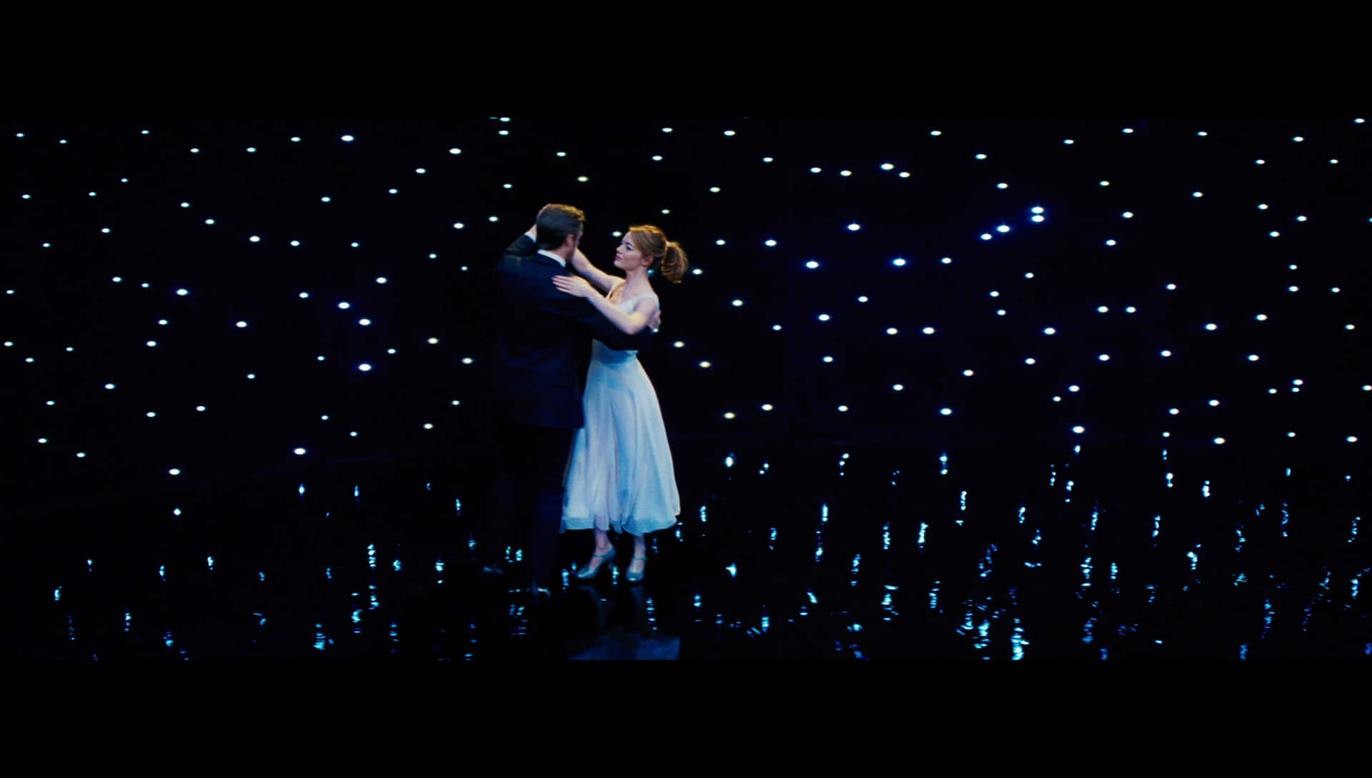 La La Land 4K 桌布 壁紙 — 一對情侶在星空般的藍色燈光展示下，於反射舞台上親密跳舞。