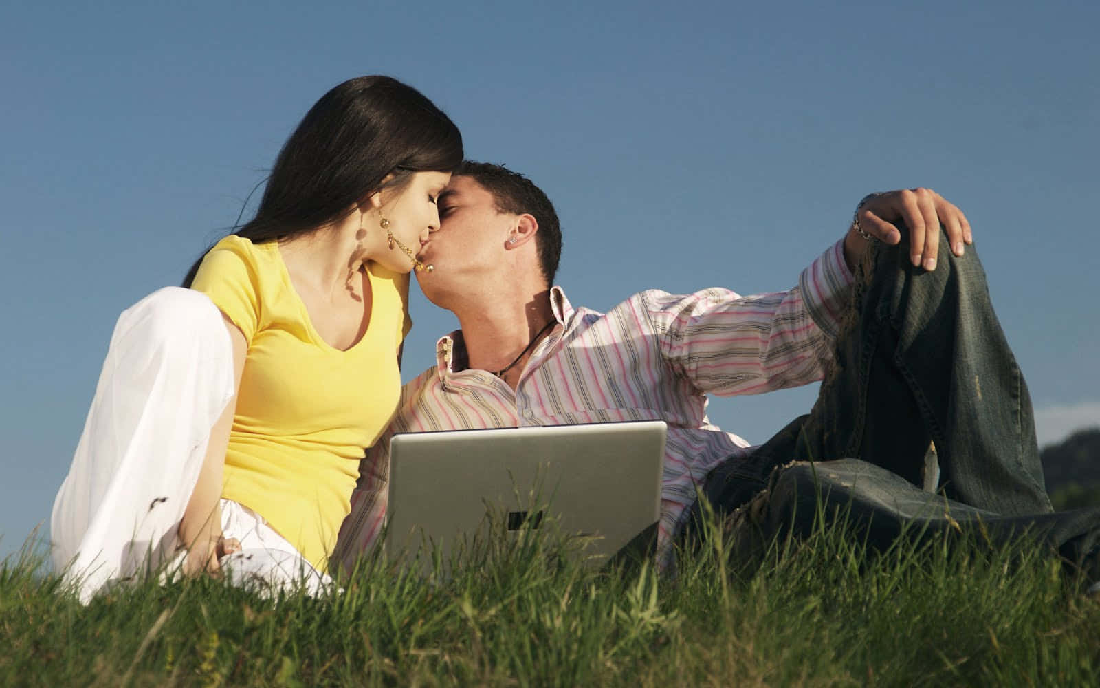 couple kiss 桌布 壁紙 — 一對情侶在晴朗藍天下躺在草地上，中間放著一台筆記型電腦，分享著溫柔的親吻。
