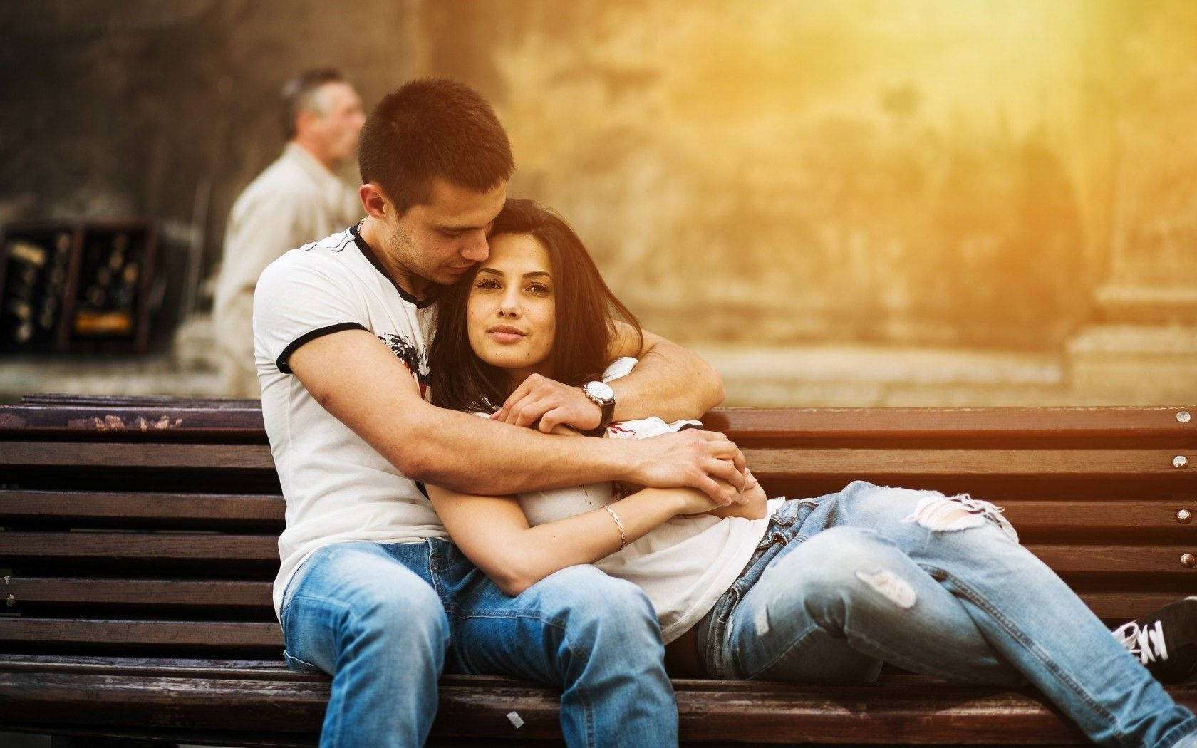 Couples 桌布 壁紙 — 一對情侶在木長椅上溫柔擁抱，金色陽光沐浴著他們親密的時刻，背景是柔和模糊的城市景致。