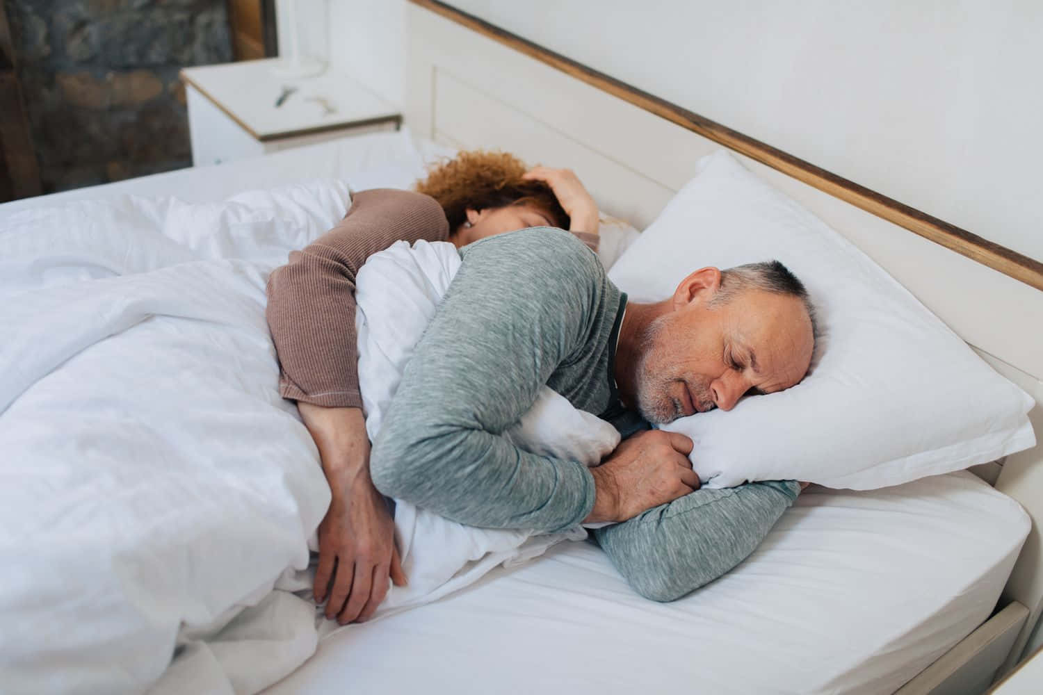People Sleeping 桌布 壁紙 — 一對情侶在柔和光線的臥室中，於白色床單和木質床頭的床上安然入睡。