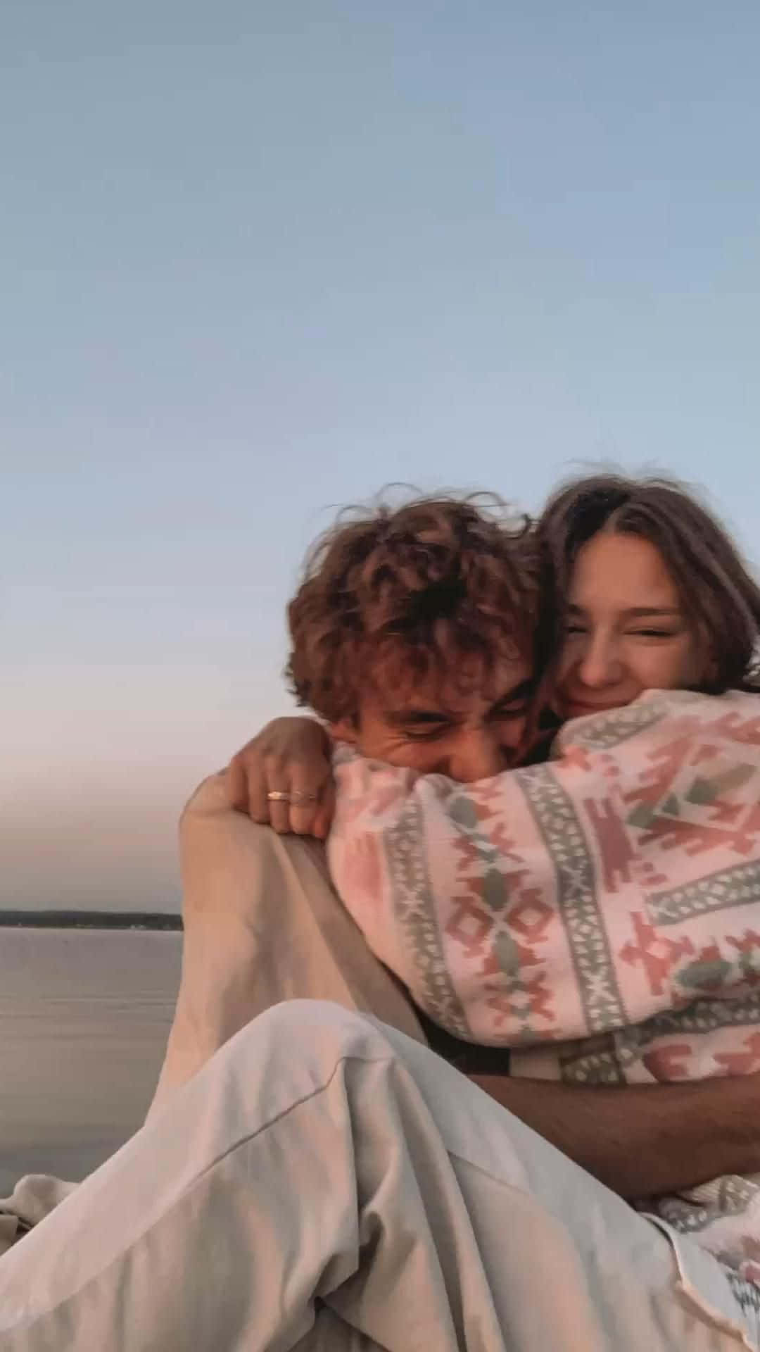 Hugging 桌布 壁紙 — 一對情侶在柔和的暮色天空下，在平靜的水邊，用一條有圖案的毯子包裹著，溫暖地擁抱。