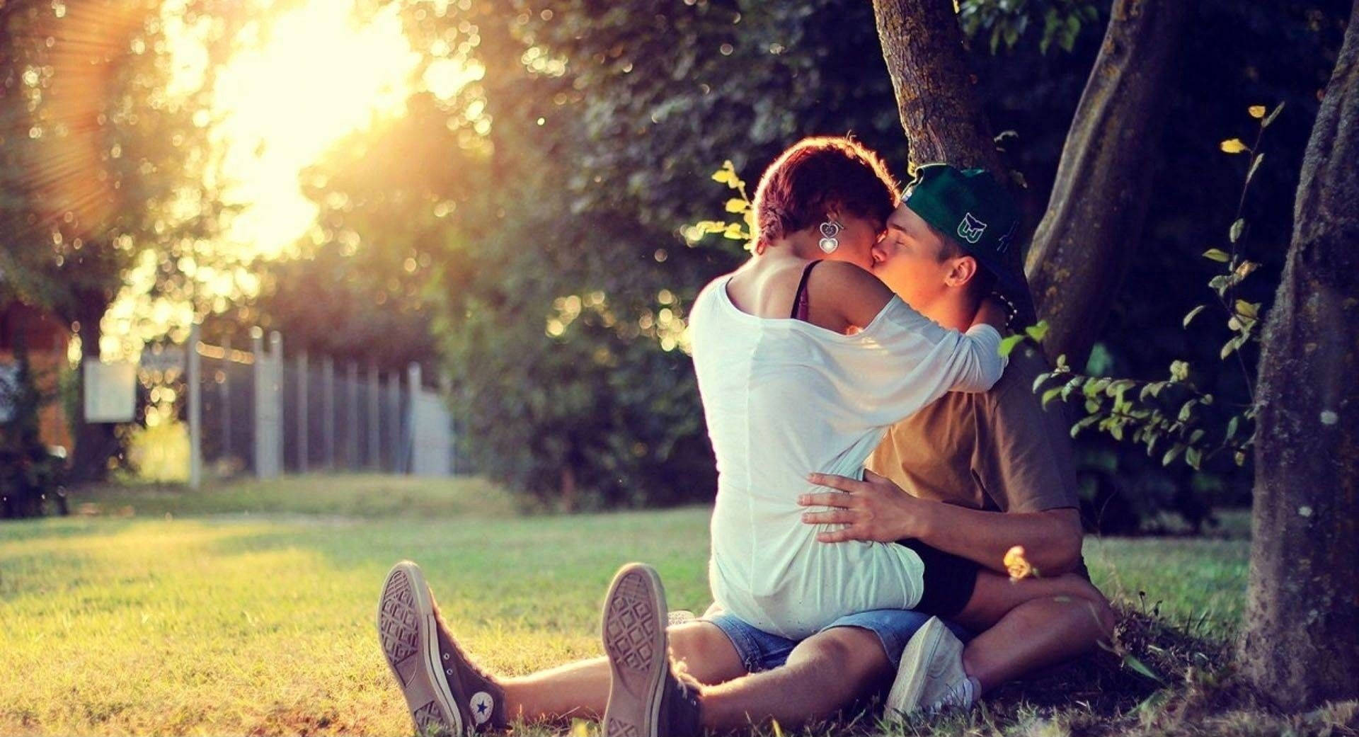couples kissing 桌布 壁紙 — 一對情侶在樹下擁抱親吻，金色陽光穿過樹葉灑落，營造出寧靜的公園氛圍。