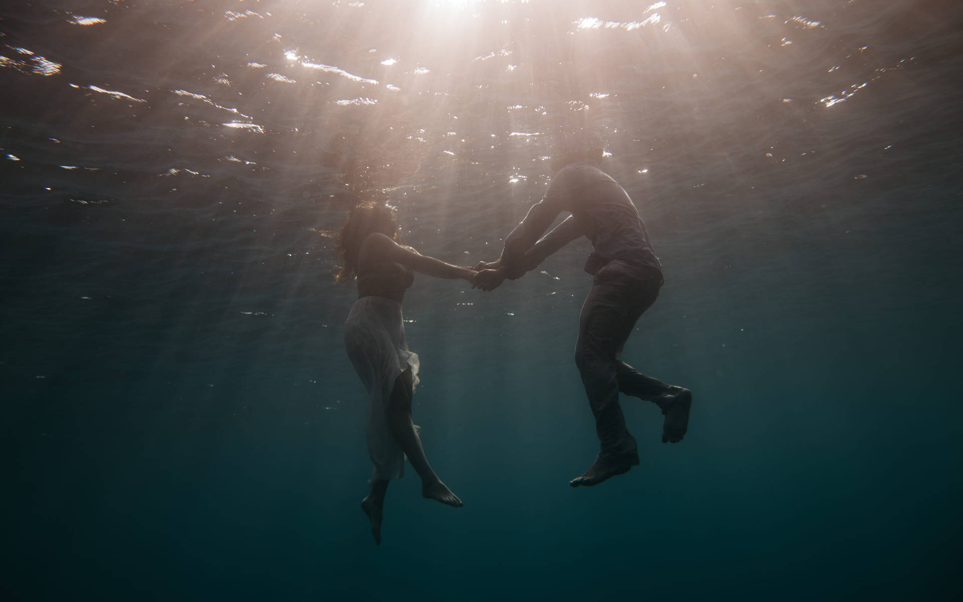 Swimming 桌布 壁紙 — 一對情侶在水底牽手，陽光從水面照射下來，營造出寧靜而浪漫的場景。