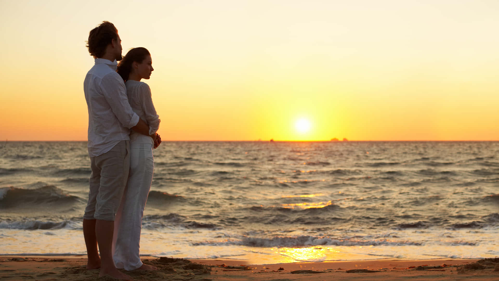 Love Couple 桌布 壁紙 — 一對情侶在沙灘上擁抱，夕陽在海面上落下，將金色的光芒灑在波浪上。