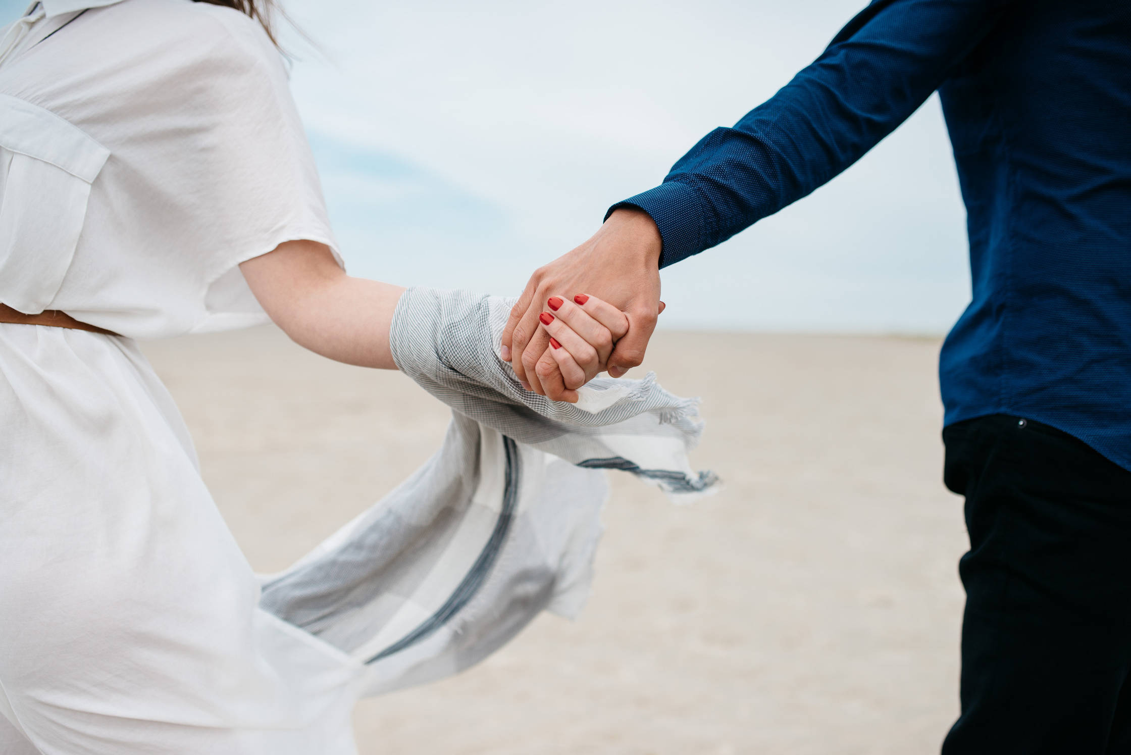 情侶 桌布 壁紙 — 一對情侶在沙灘上牽手漫步，風吹動他們的衣服。