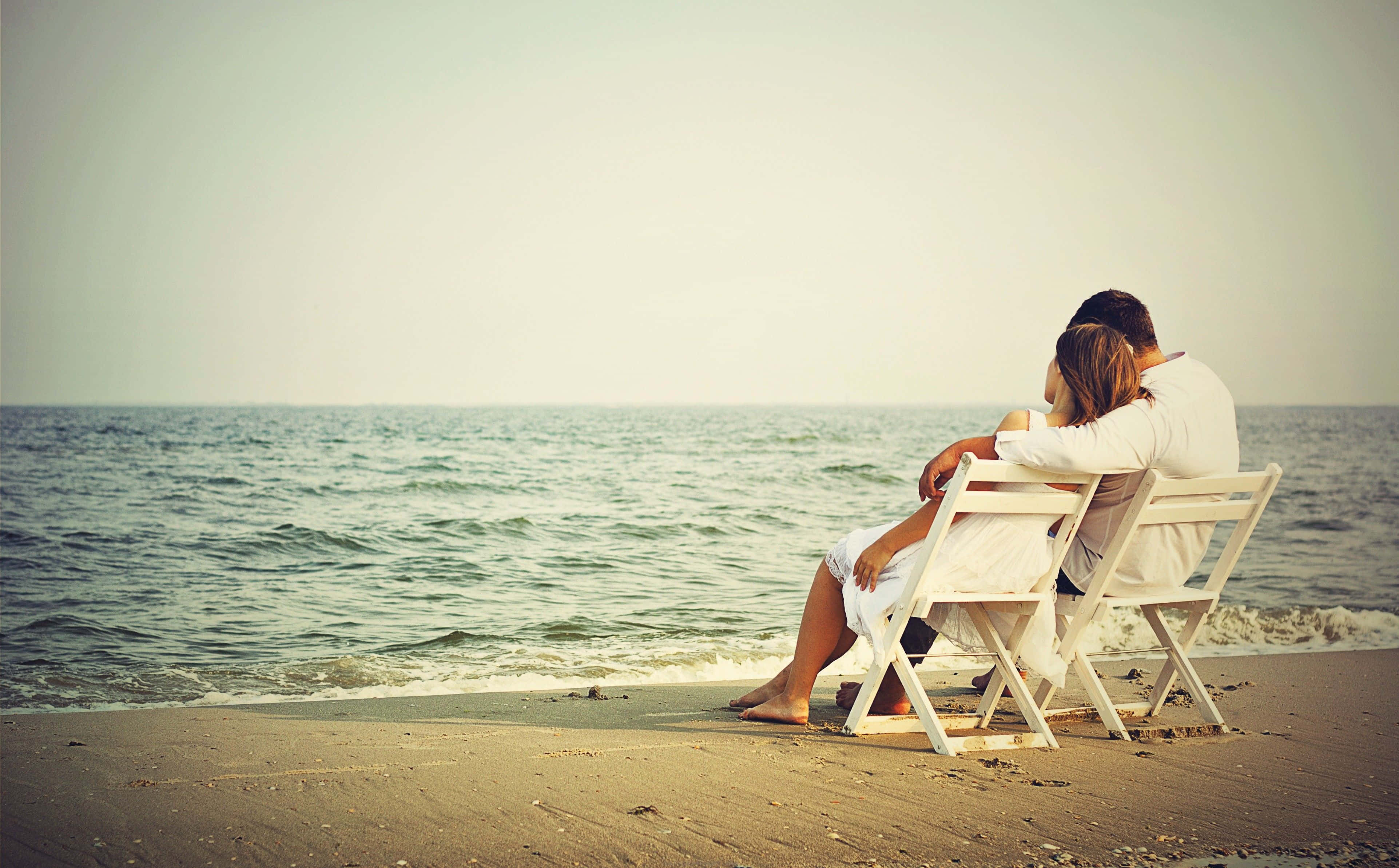 Beach Lovers 桌布 壁紙 — 一對情侶在沙灘上緊靠著白色椅子坐著，凝視著柔和天空下平靜的海洋。