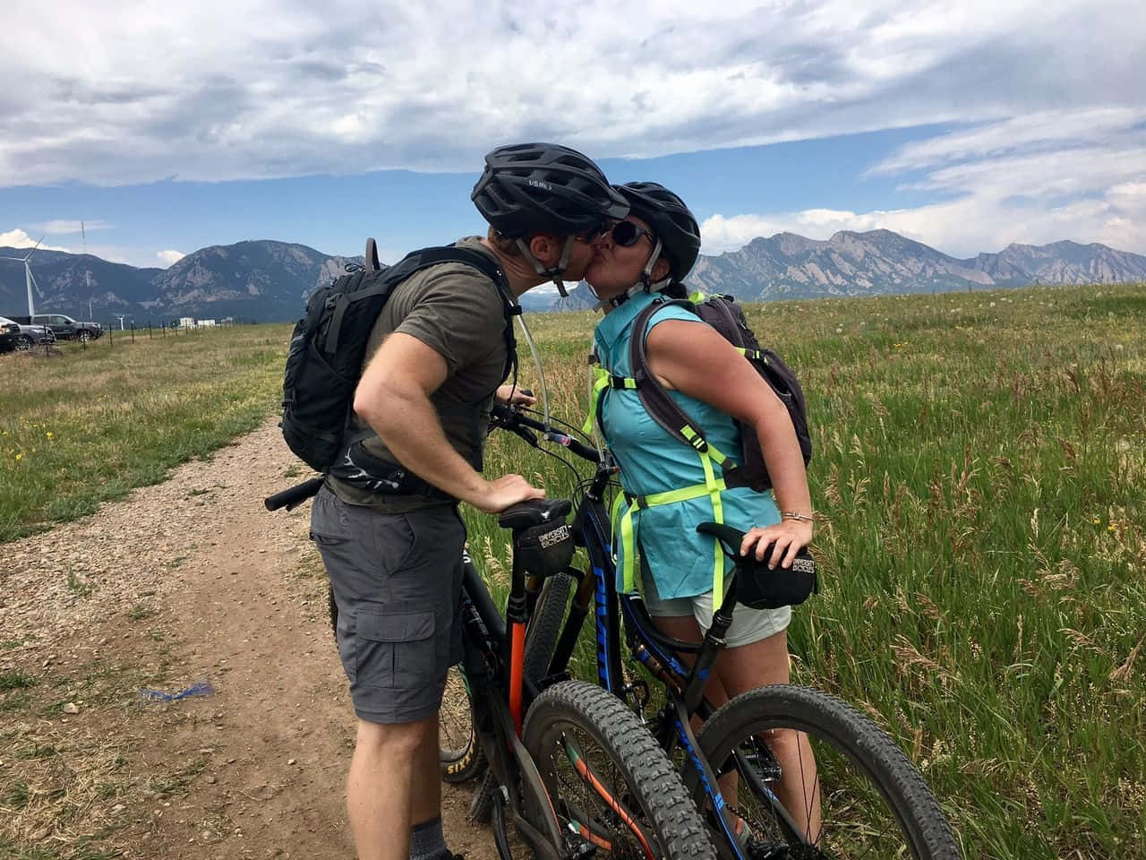bike riding 桌布 壁紙 — 一對情侶在泥濘小徑上與登山自行車站在一起接吻，周圍是草地和遠山，天空多雲。