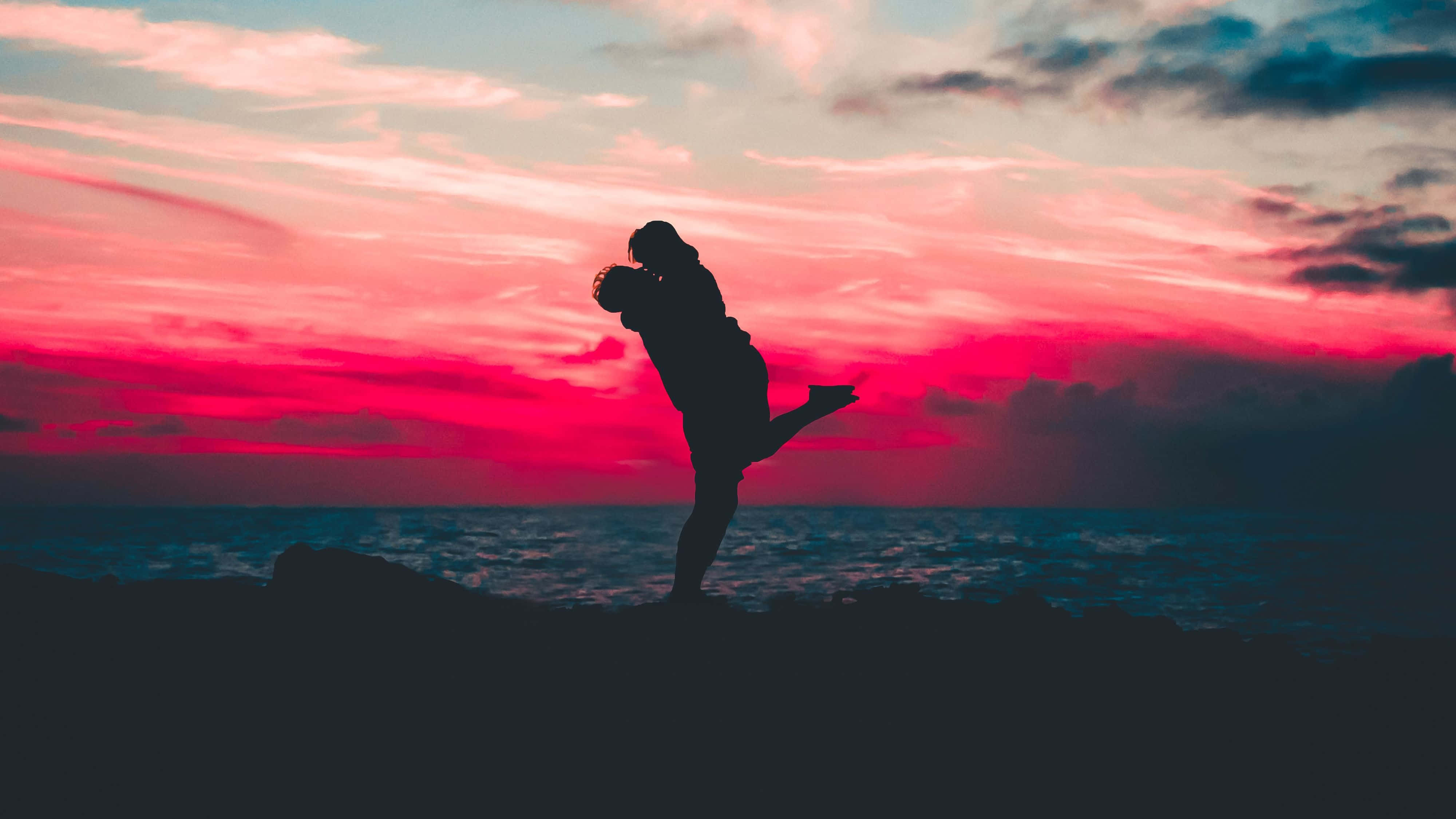 couple love 桌布 壁紙 — 一對情侶在海洋上空火熱的粉紅色和橙色日落中擁抱成剪影。