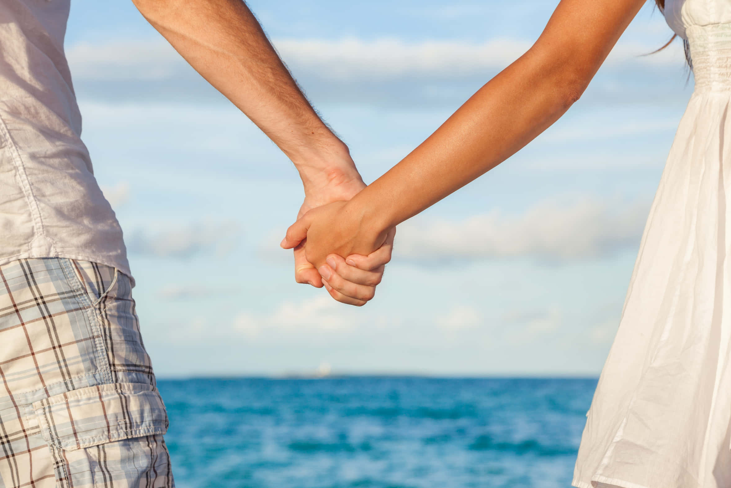Love Couple 桌布 壁紙 — 一對情侶在海洋邊牽手，天空柔和藍色，雲朵散佈。
