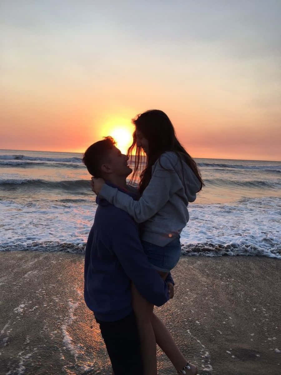 Cute Boyfriend 桌布 壁紙 — 一對情侶在海灘上以剪影擁抱，夕陽以溫暖的橙色和粉紅色調在海面上落下。