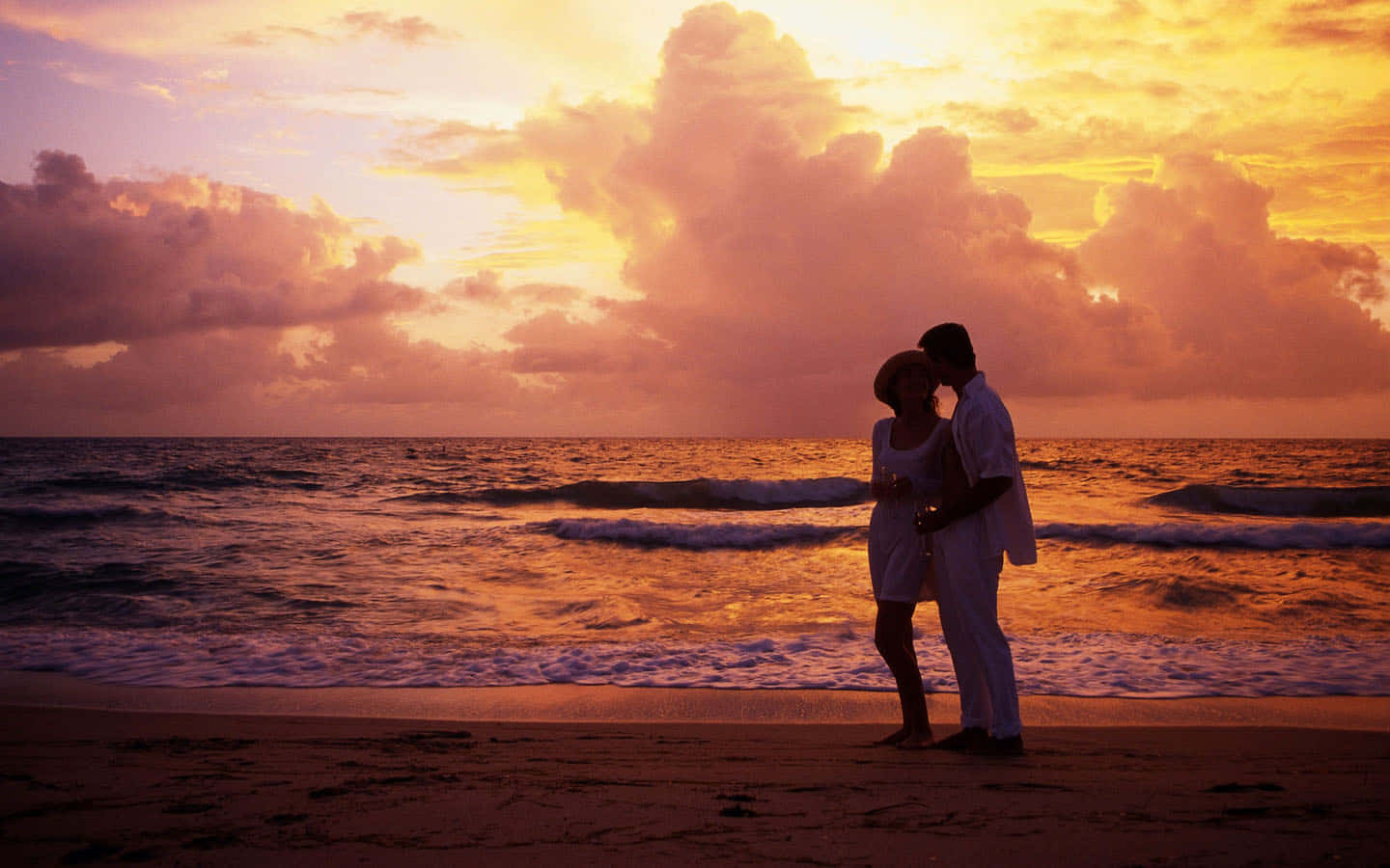 Romantic Love 桌布 壁紙 — 一對情侶在海灘上以剪影擁抱，金色夕陽的光芒以溫暖的色調沐浴著海洋和雲彩。