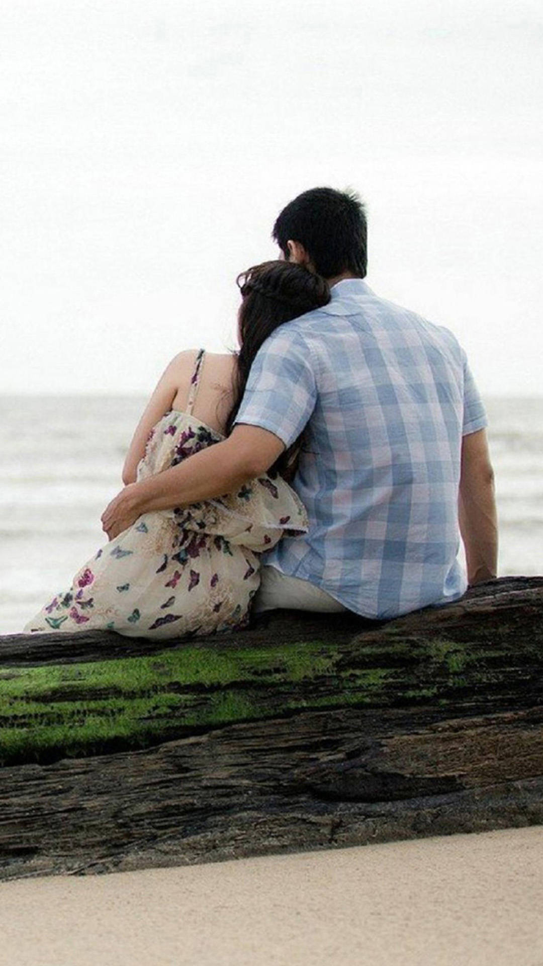 Couple 桌布 壁紙 — 一對情侶在海灘上坐在長滿苔蘚的木頭上，從後面擁抱，凝視著多雲天空下平靜的海洋。