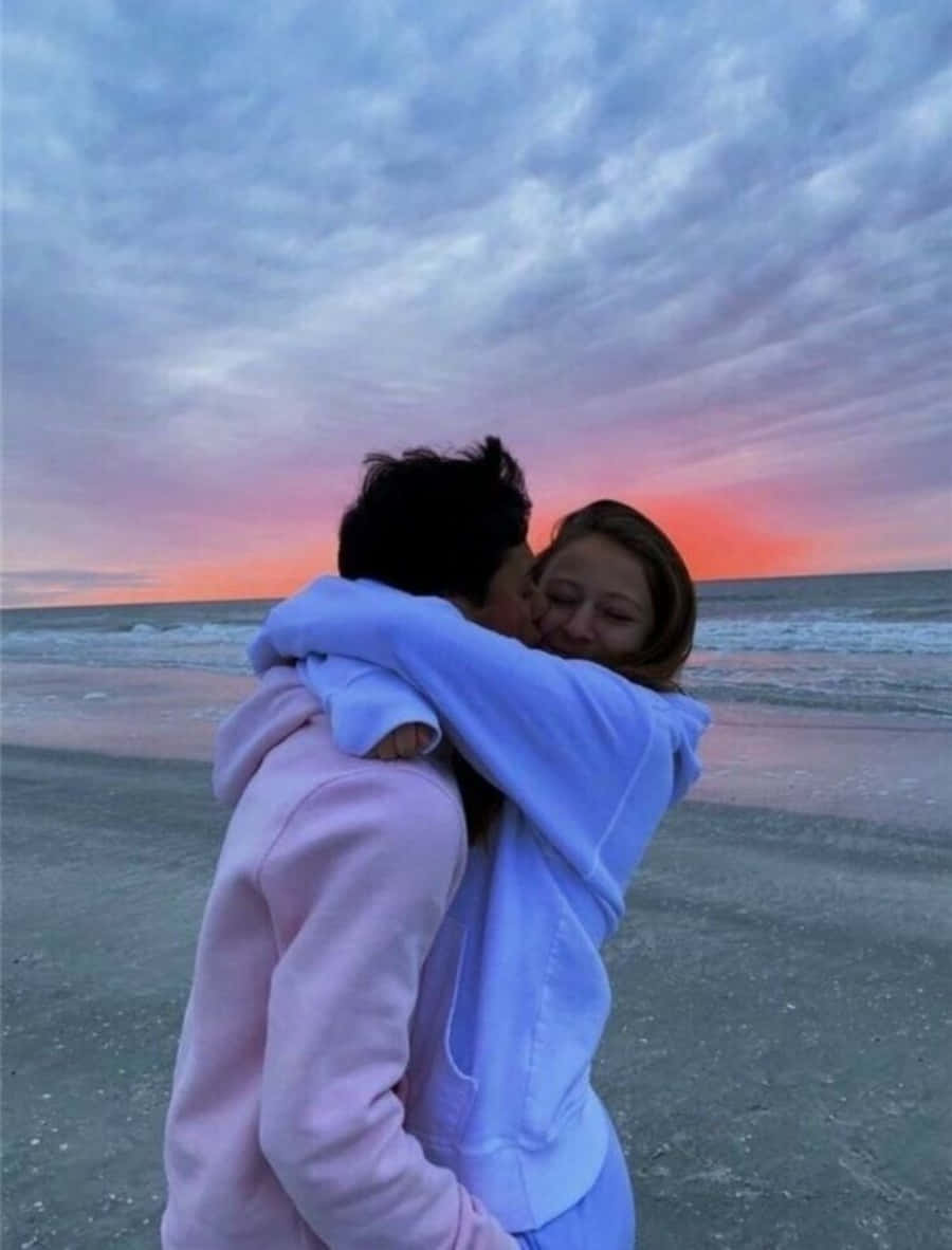 Hug 桌布 壁紙 — 一對情侶在海灘上擁抱，天空在日落時呈現粉紅色和橙色。