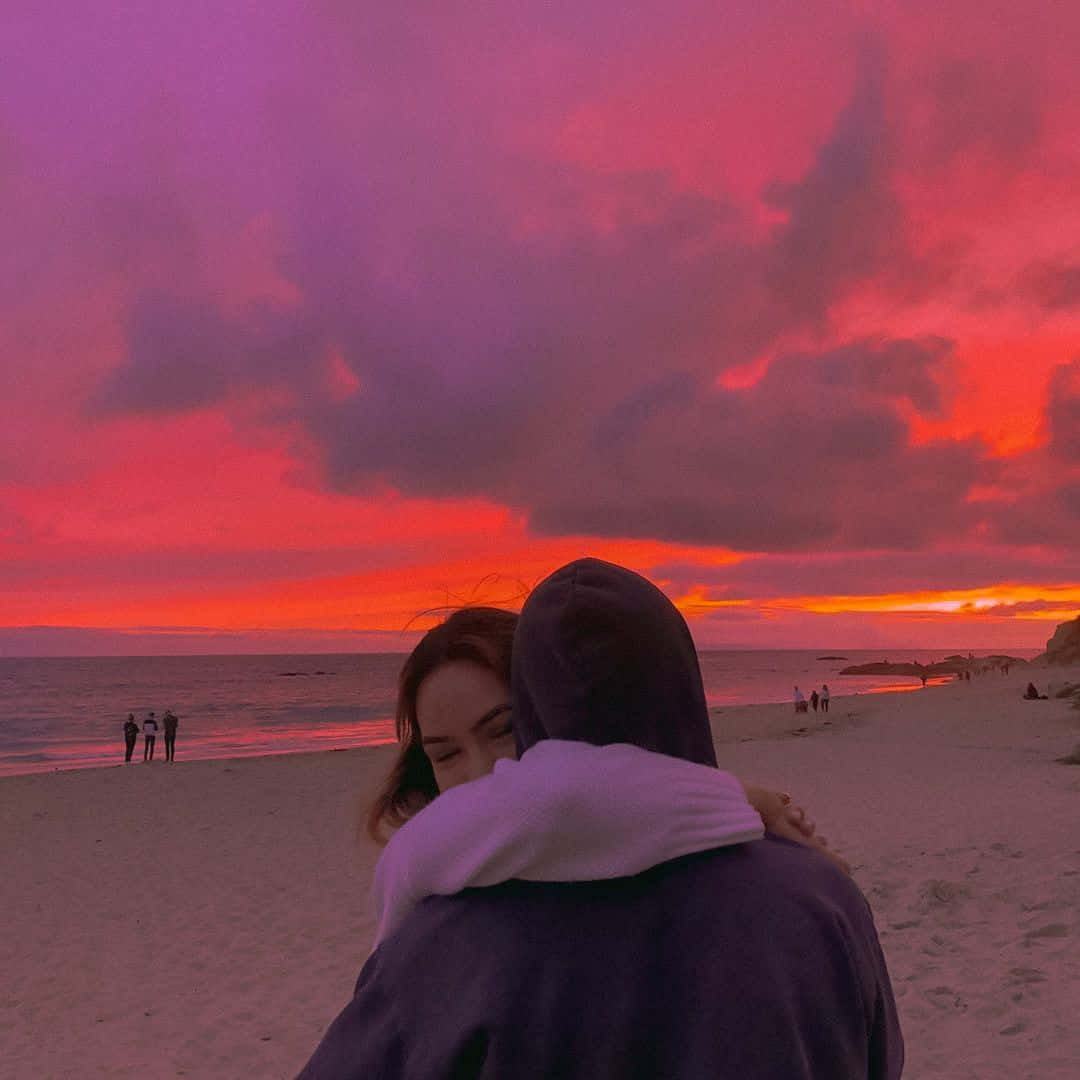 Boyfriend And Girlfriend 桌布 壁紙 — 一對情侶在海灘上擁抱，火熱的粉紅色和橙色夕陽描繪了天空和海洋。