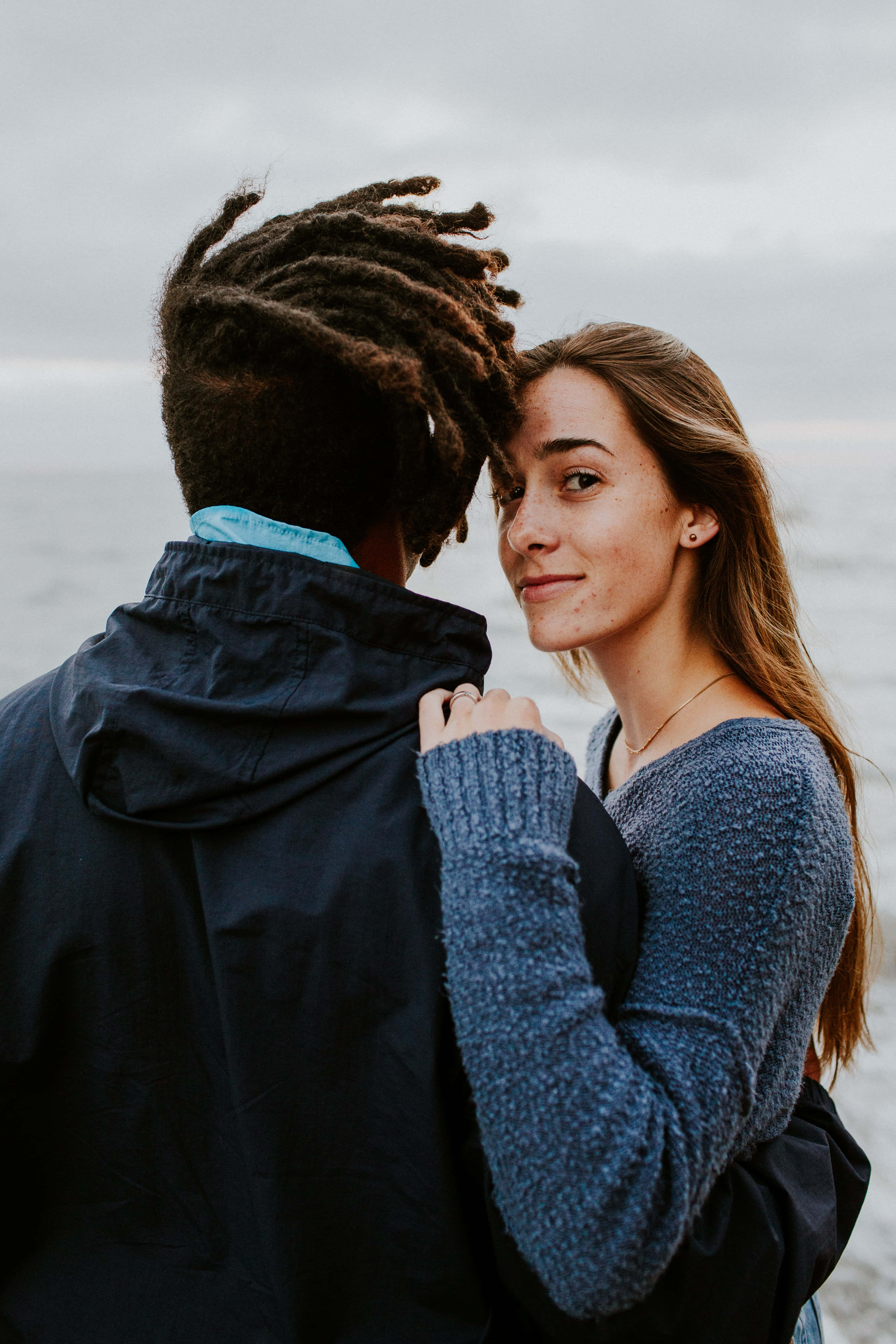 Interracial 桌布 壁紙 — 一對情侶在海邊擁抱，女士在男士的肩膀上微笑，背景是陰天。
