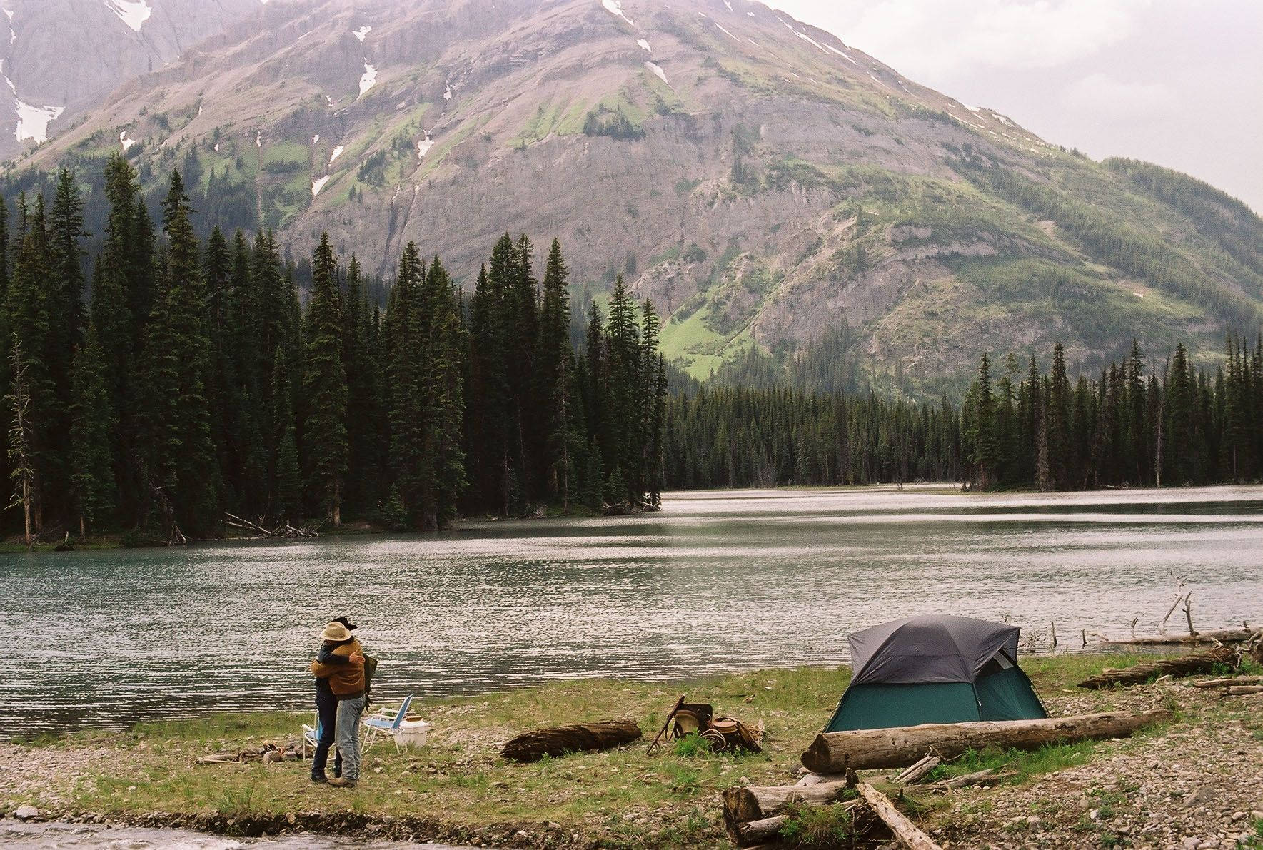 brokeback mountain 桌布 壁紙 — 一對情侶在湖邊營地擁抱，營地有一個綠色帳篷，背景是高聳的山脈和茂密的松樹林。
