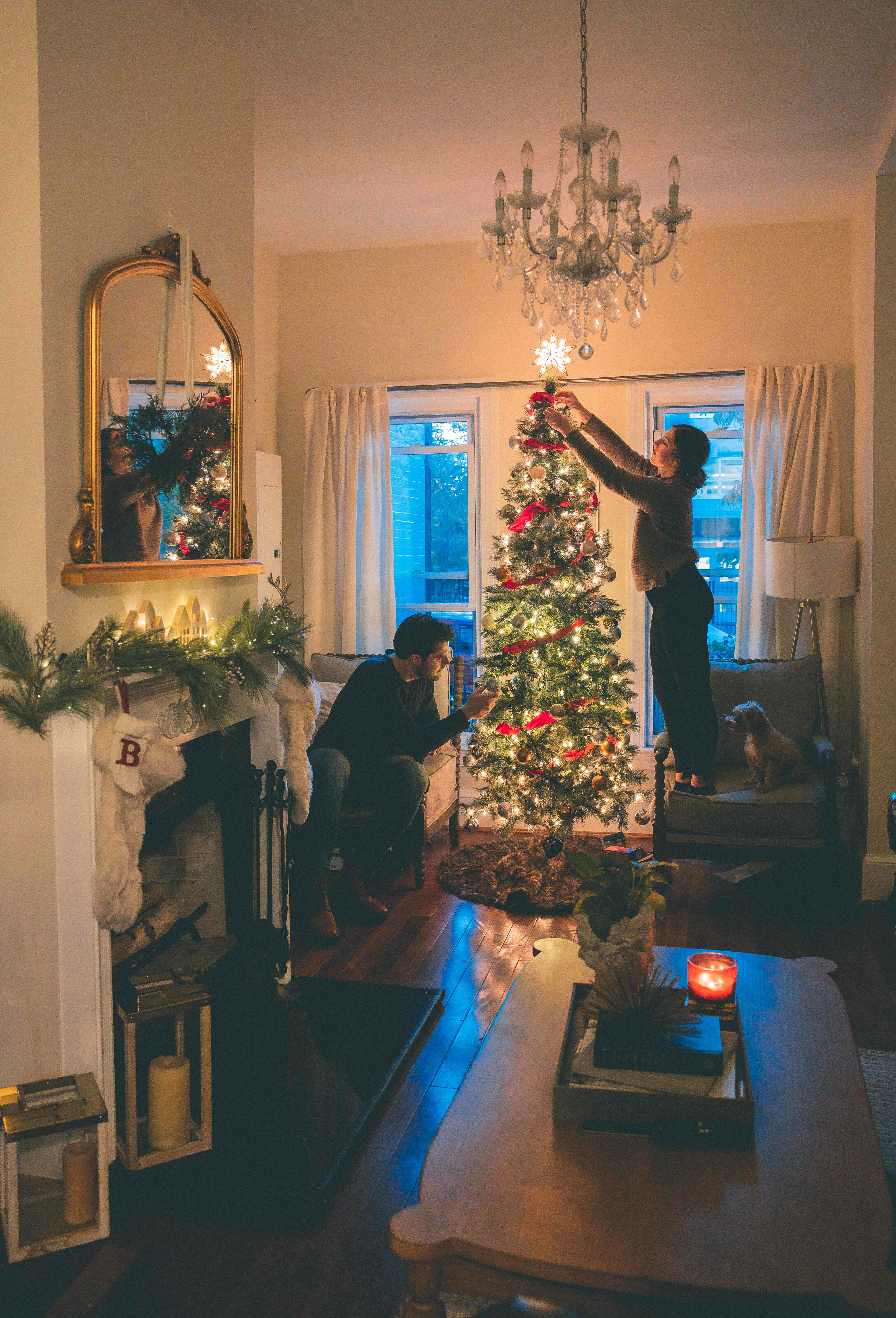 Christmas Scene 桌布 壁紙 — 一對情侶在溫暖照明的客廳中裝飾發光的聖誕樹，客廳有吊燈和壁爐。