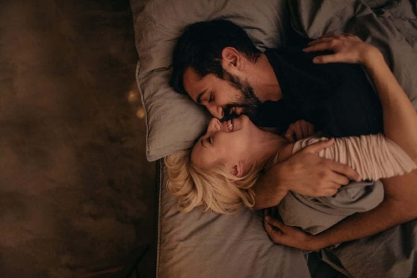 kissing in bed 桌布 壁紙 — 一對情侶在溫暖的環境燈光下，在柔軟灰色床單的床上溫柔擁抱。