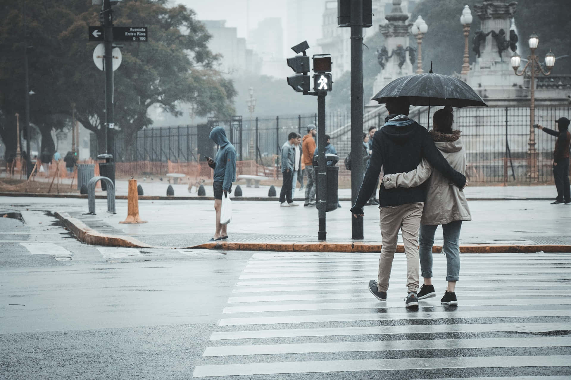 情侶 桌布 壁紙 — 一對情侶在灰色雨中共用一把傘，穿過濕漉漉的城市街道，背景有模糊的行人和一座紀念碑。