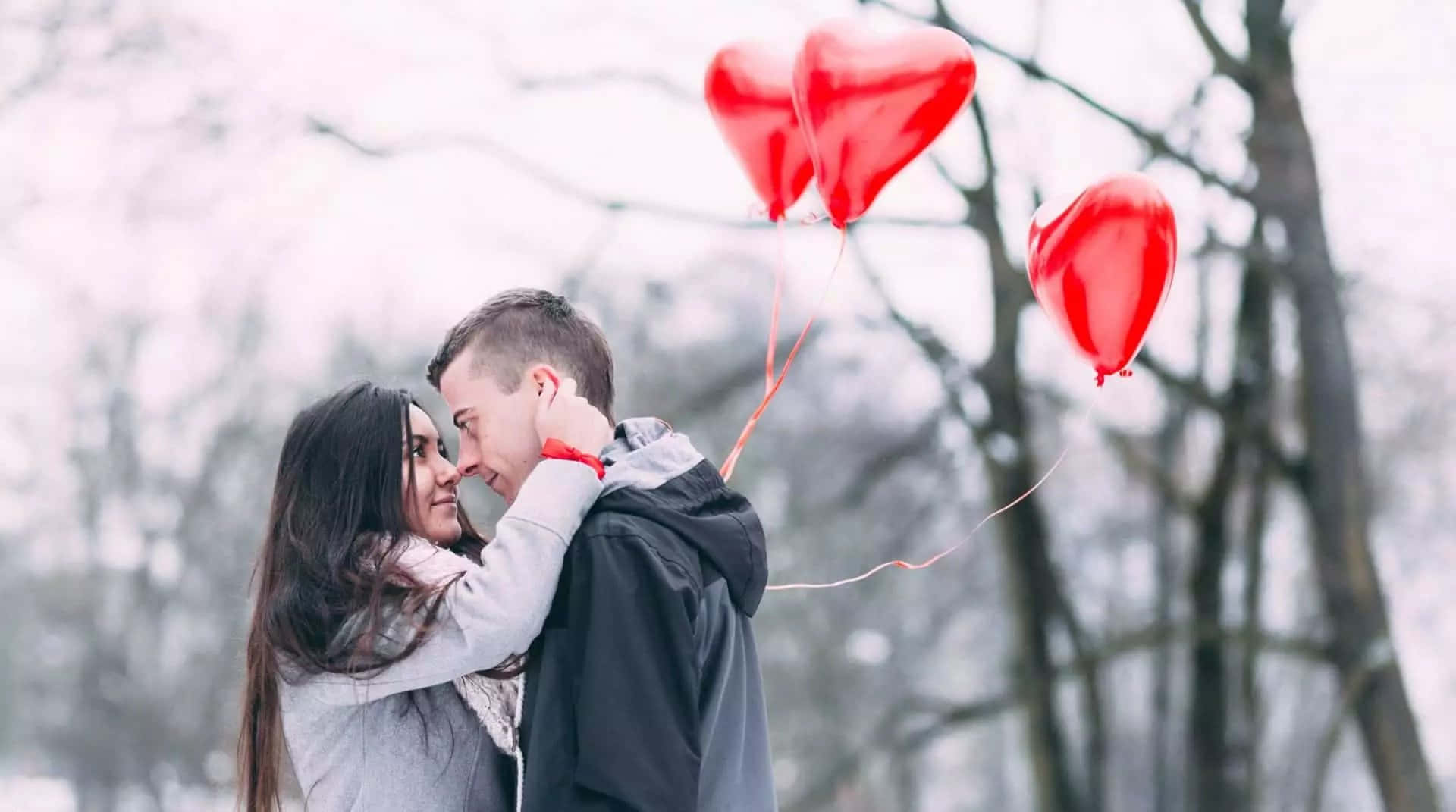 Falling In Love 桌布 壁紙 — 一對情侶在白雪覆蓋的公園中溫柔擁抱，三顆紅色心形氣球在光禿的冬季樹木襯托下向上飄升。