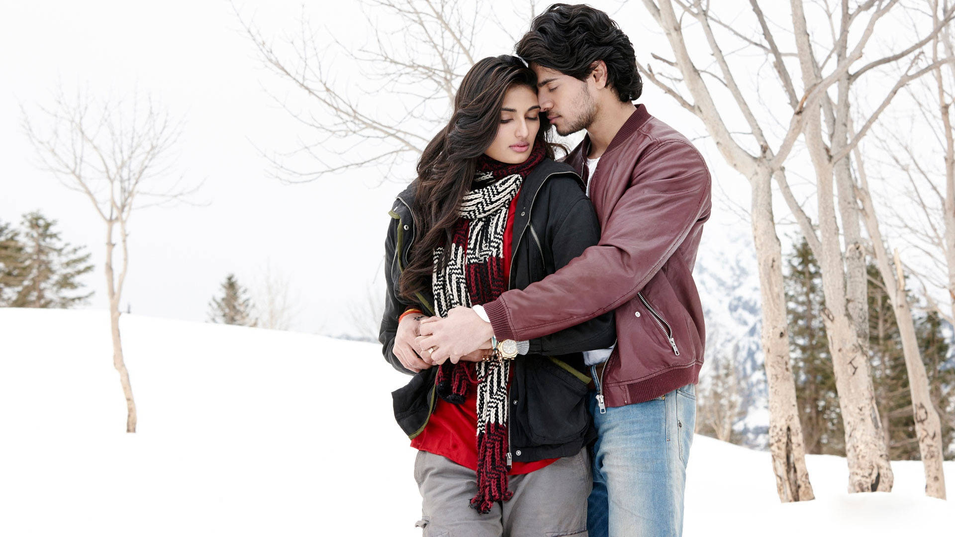 Bollywood HD 桌布 壁紙 — 一對情侶在白雪覆蓋的冬季森林中溫柔擁抱，背景有光禿的樹木和遠處的山脈，天空多雲。