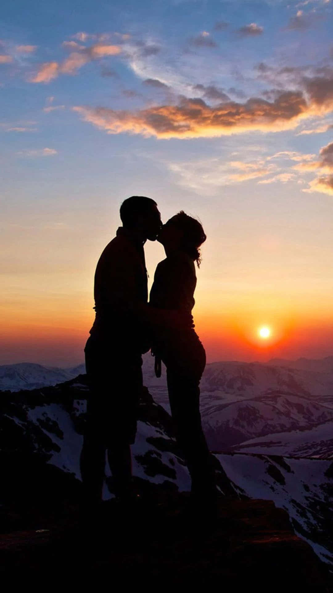 Romantic Love 桌布 壁紙 — 一對情侶在白雪覆蓋的山峰頂上以剪影接吻，夕陽在他們身後落下，將溫暖的橙色和粉紅色調灑滿天空。