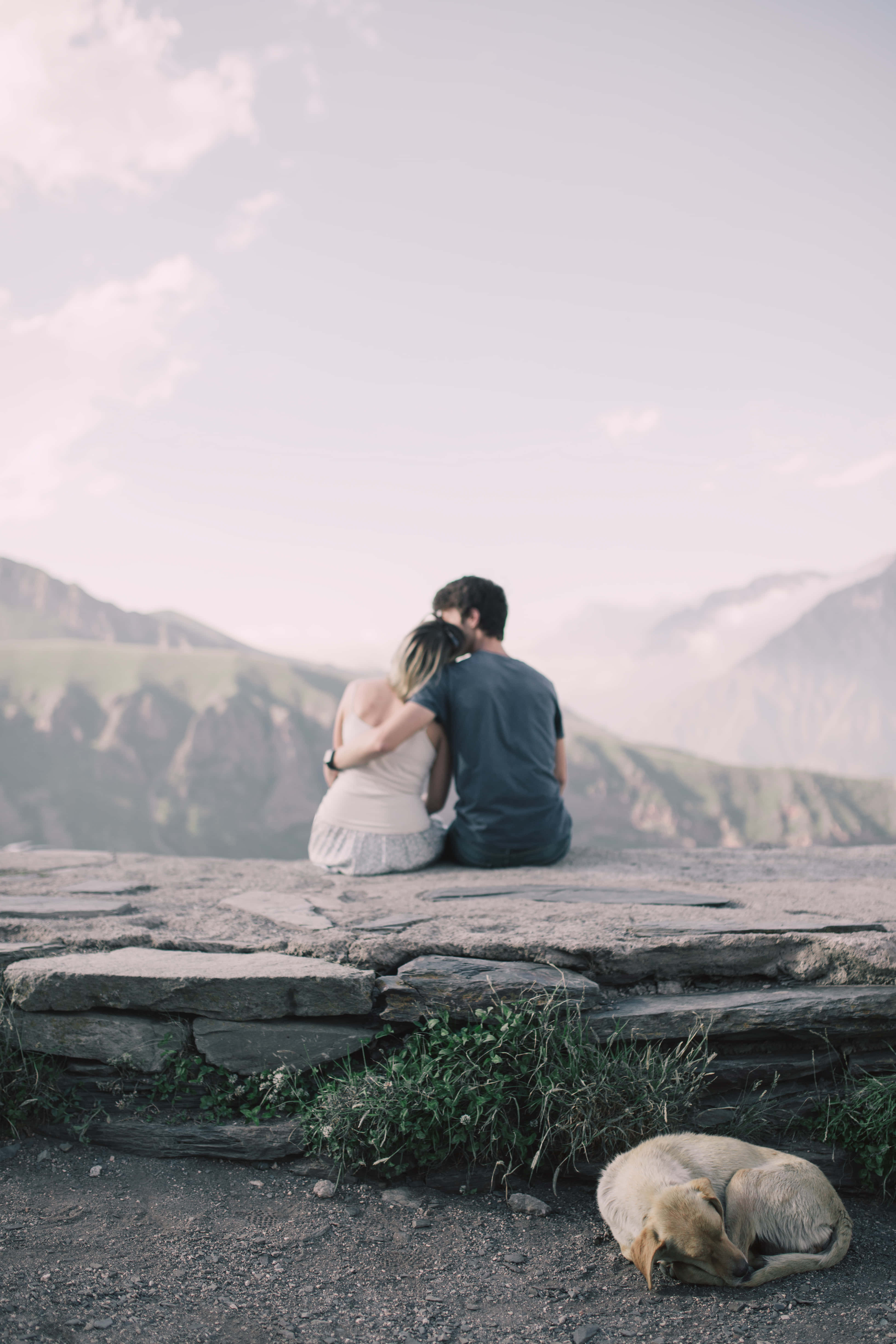 Lovers 桌布 壁紙 — 一對情侶在石崖上擁抱，俯瞰煙霧繚繞的山脈，附近有一隻正在睡覺的狗。